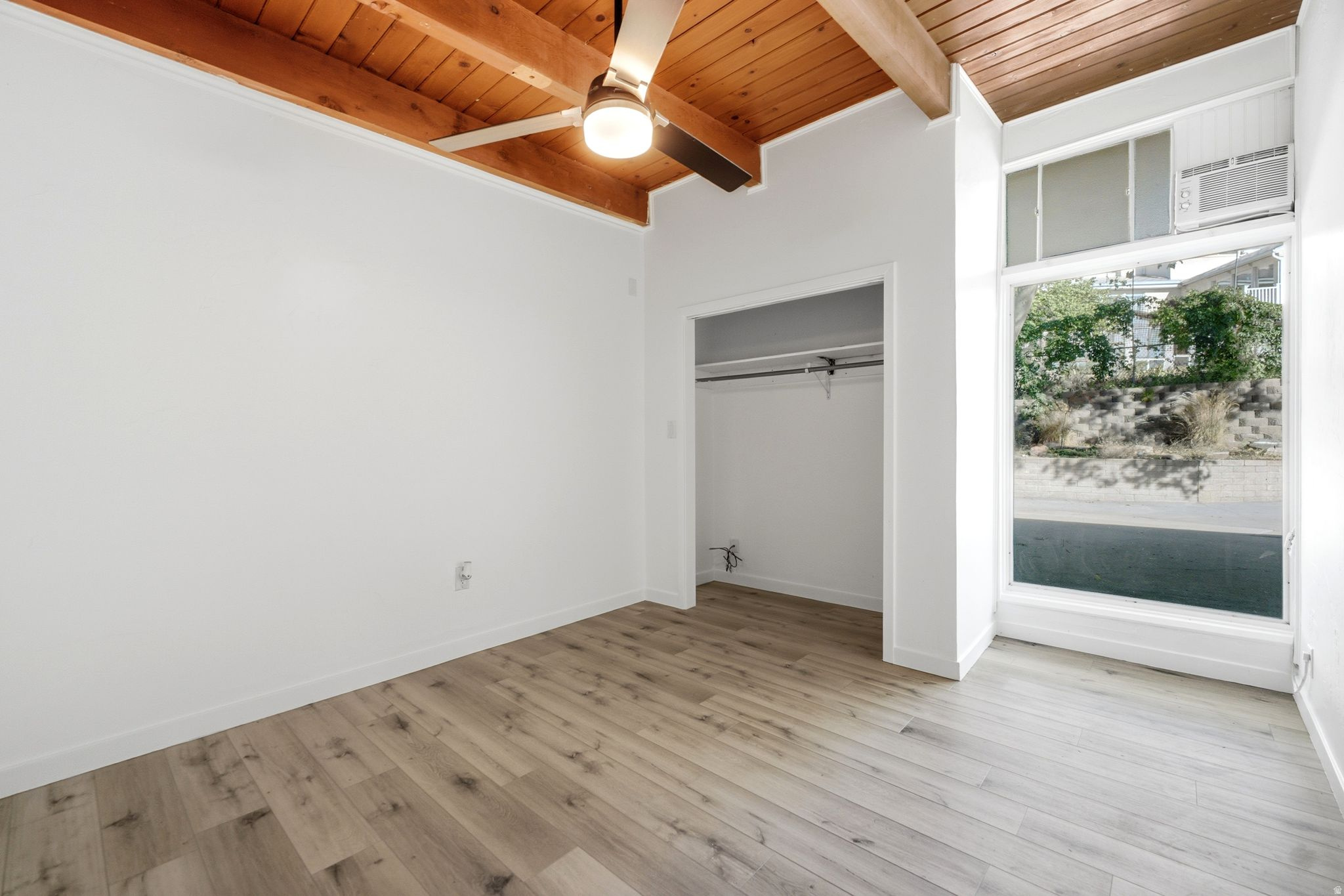 Bright corner bedroom with wood-finish flooring, exposed beam ceiling with recessed lighting, large windows with natural light, and a spacious closet with sliding door