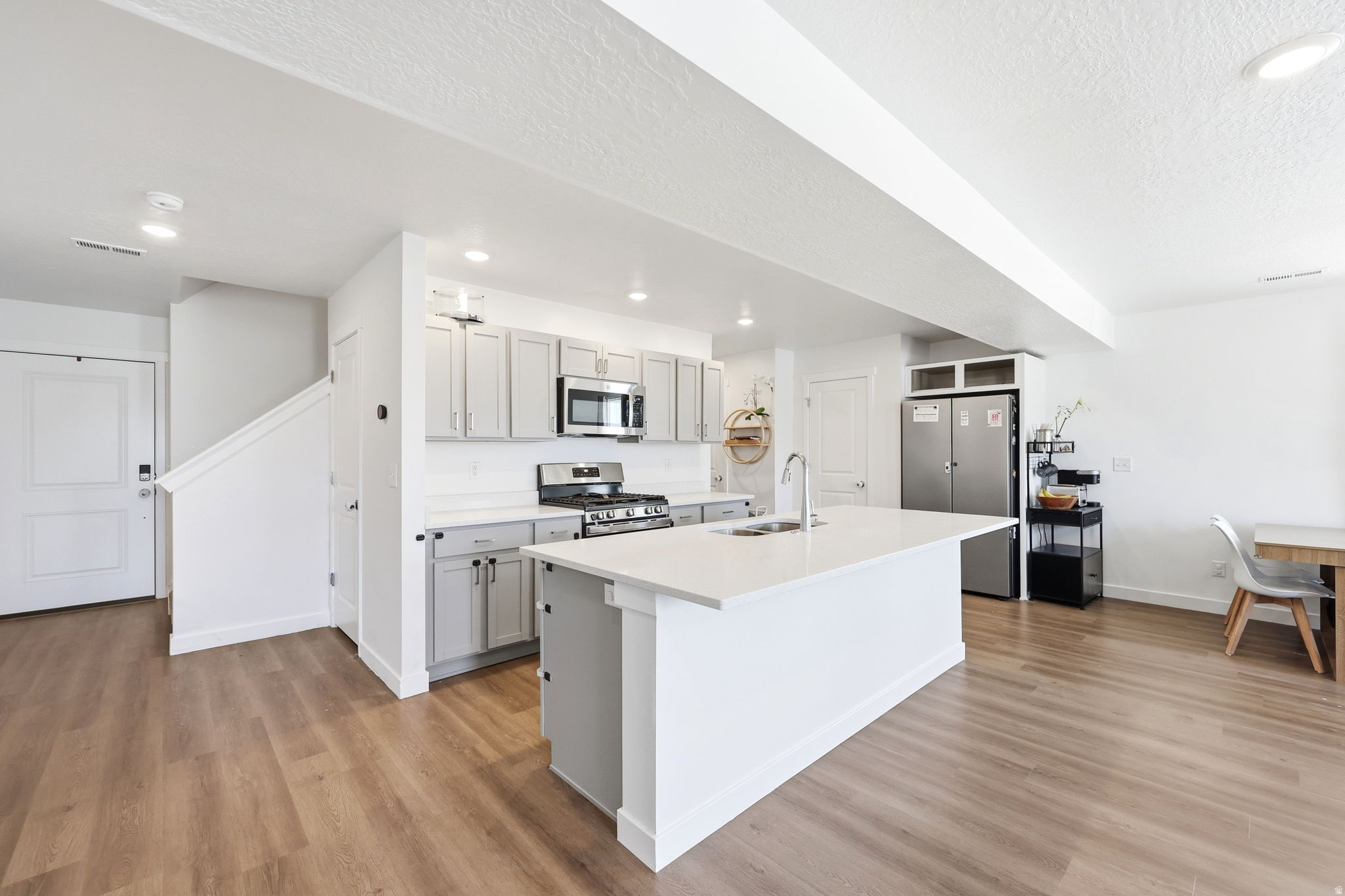 Spacious kitchen featuring an expansive central island with a sink, light gray cabinetry, stainless steel appliances, white countertops, and wood-finish flooring