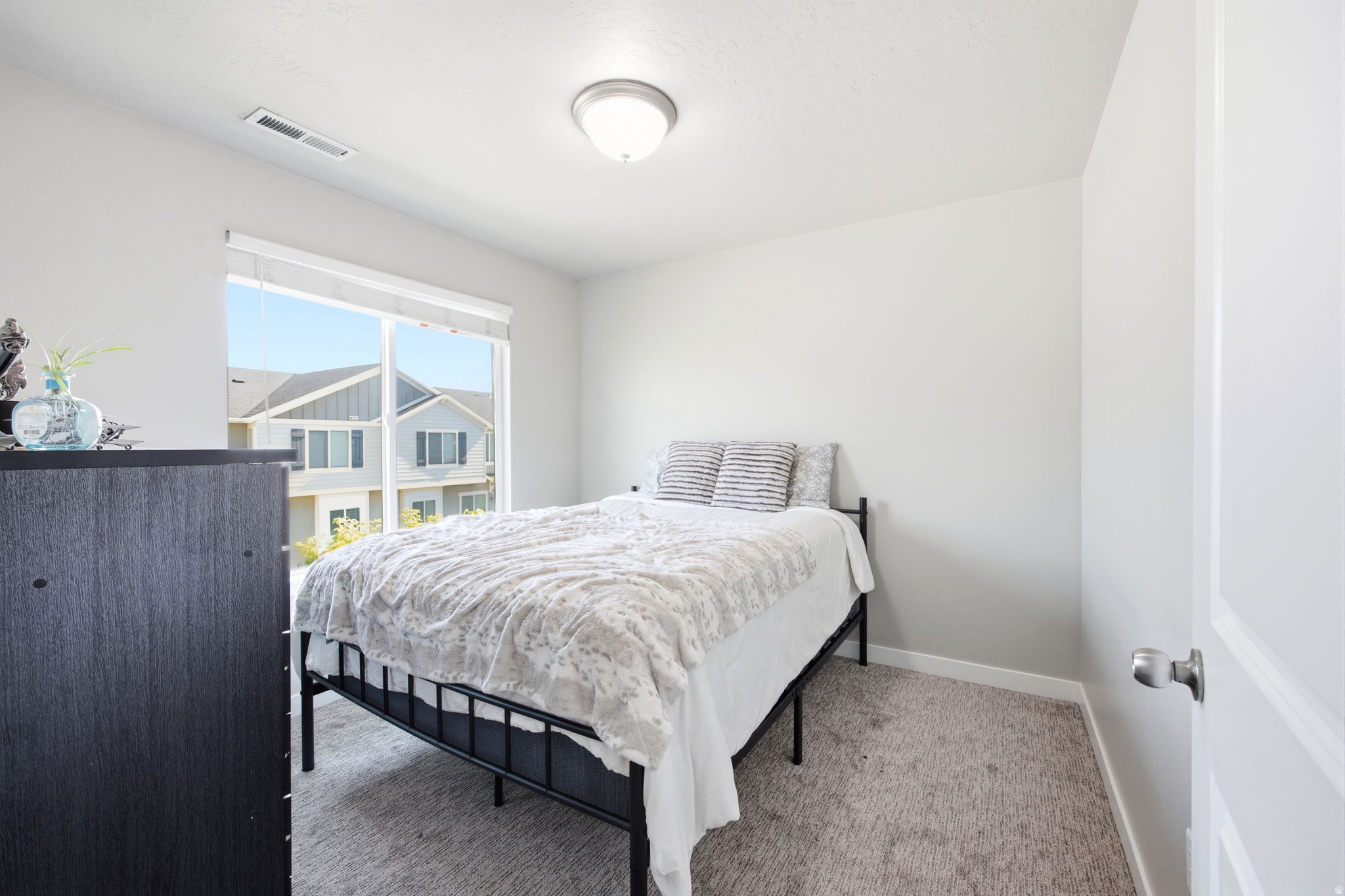 Bright bedroom featuring a large window with roller shades, light gray wall paint, and gray carpet flooring