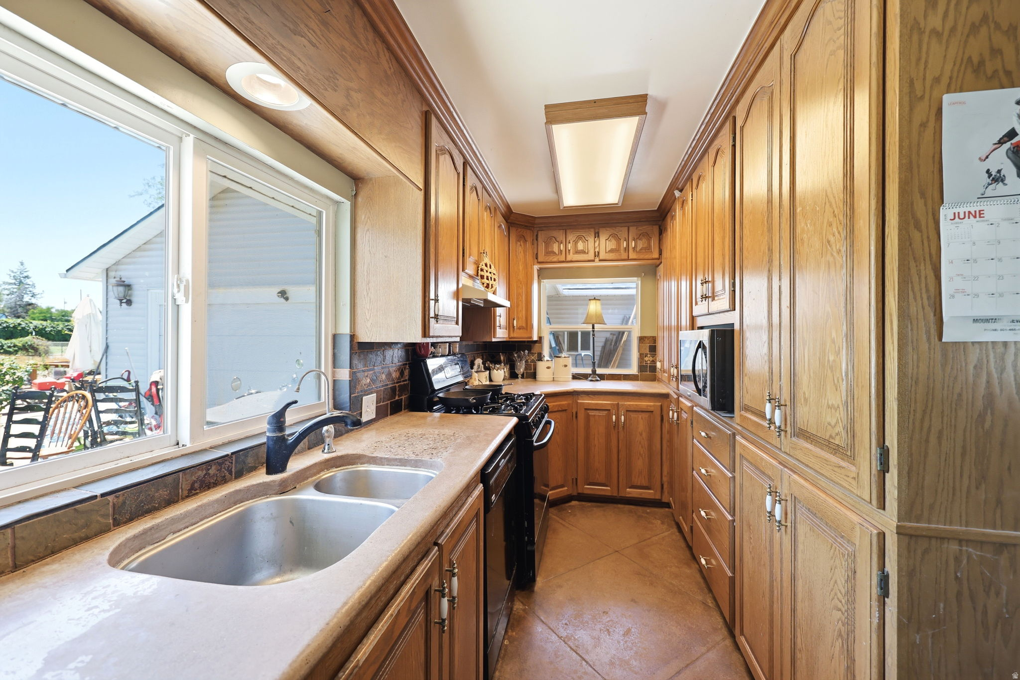 Galley kitchen featuring wood cabinetry, stone-finish countertops, stainless steel double-basin sink with a matte black faucet, and tiled backsplash