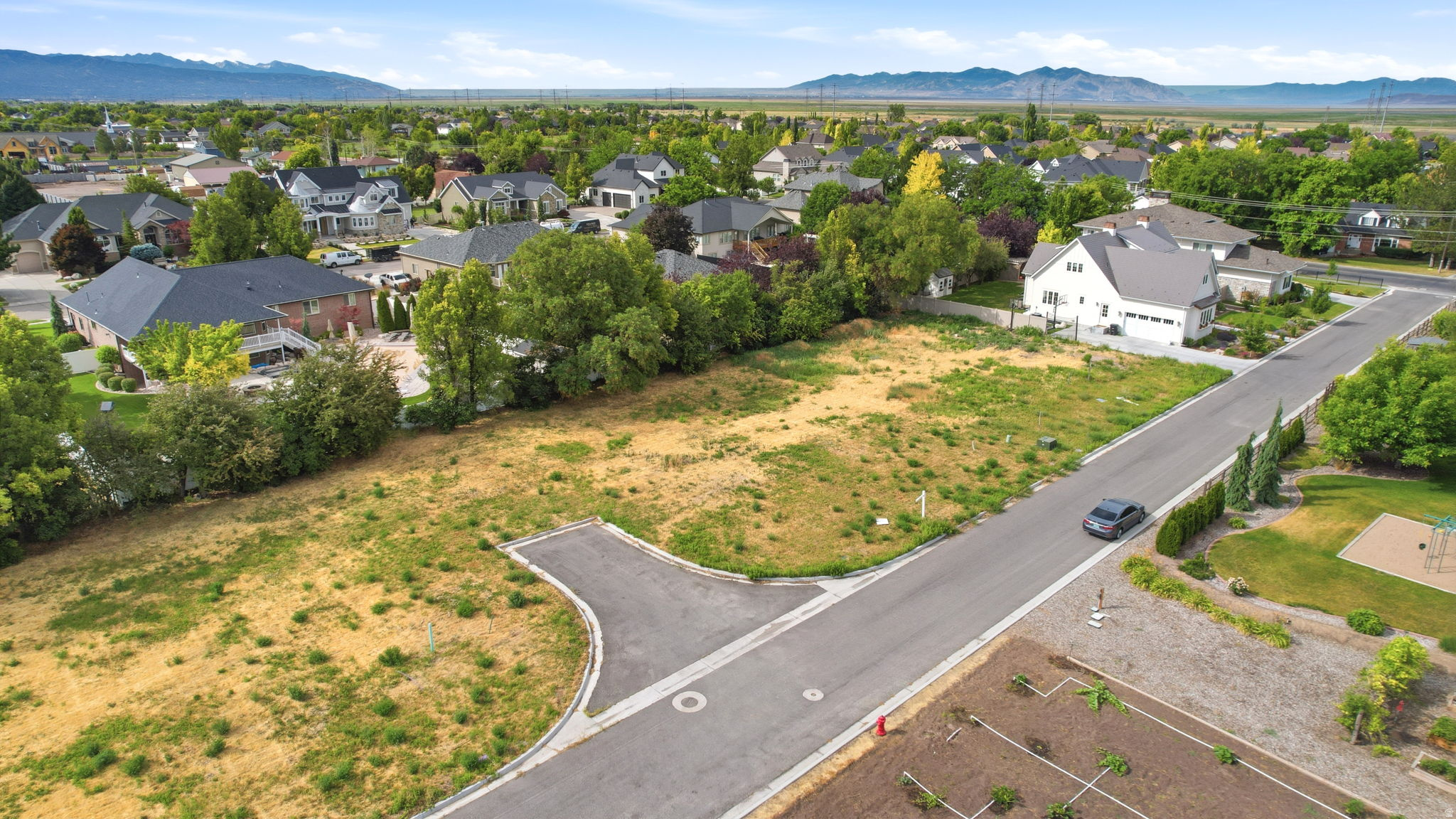 Expansive undeveloped lot featuring a paved road frontage, established surrounding residential properties, mature trees, and distant mountain backdrops