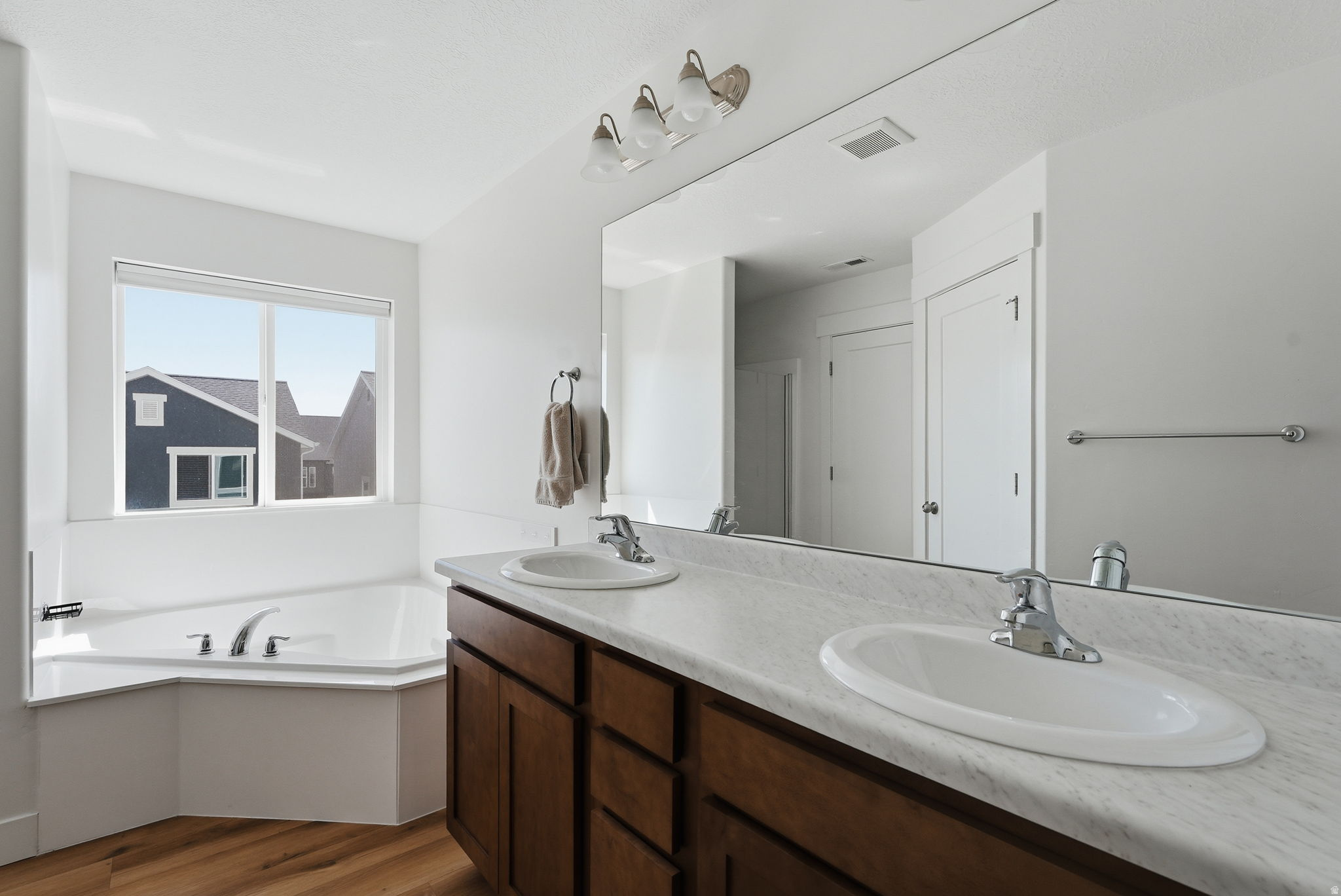 Bathroom featuring a corner soaking tub, a dual vanity with a large mirror, wood-finish flooring, and white walls