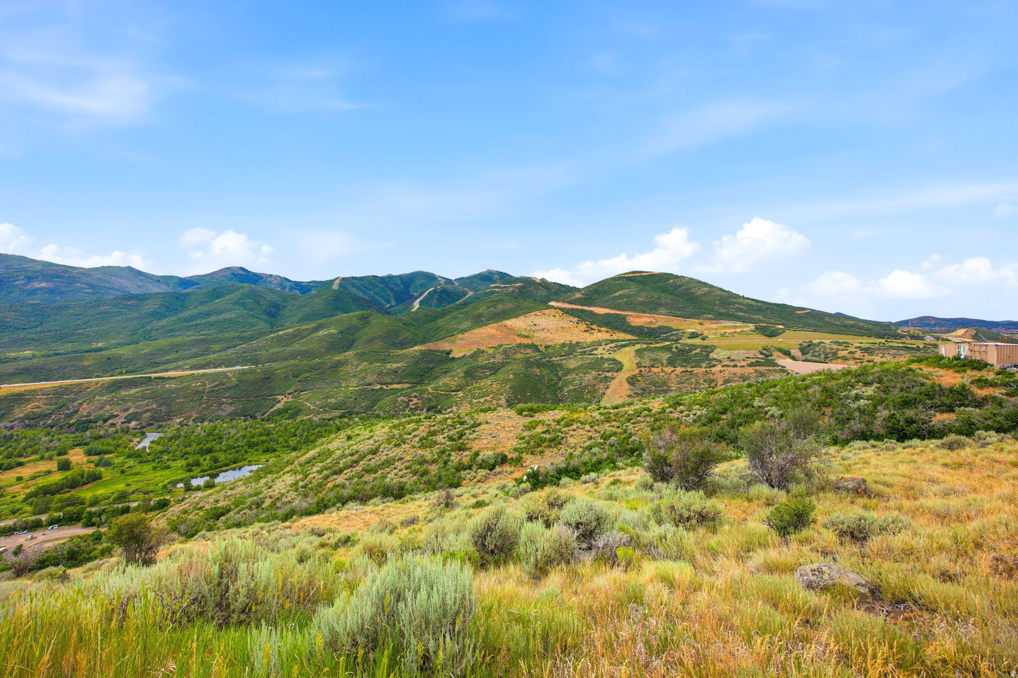 Expansive mountain landscape featuring varied green and golden-toned foliage, a winding waterway, and distant structures under a clear sky