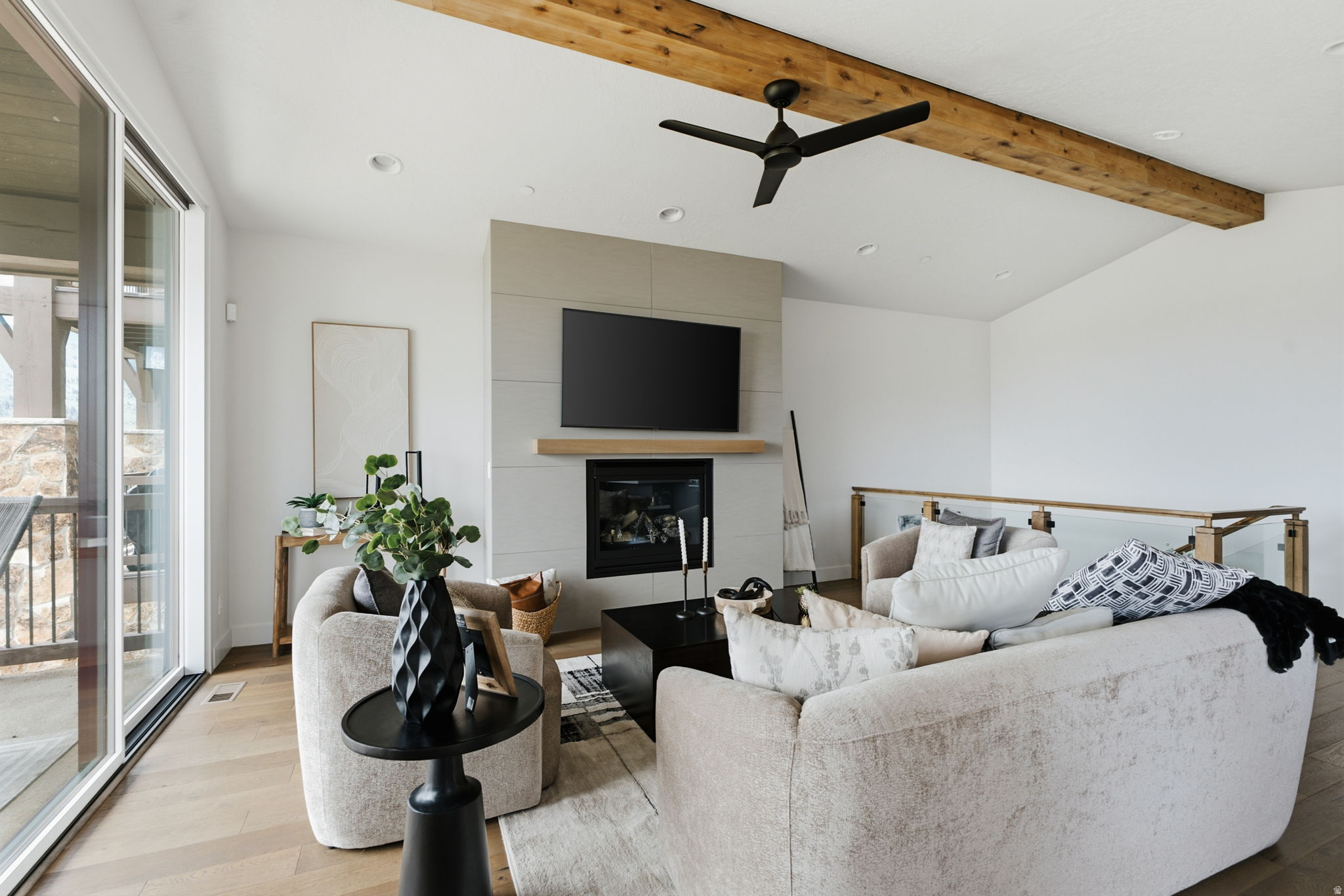 Spacious living area featuring a vaulted ceiling with exposed wood beams, a contemporary fireplace with a mounted television, wood-finish flooring, and large sliding glass doors leading to an outdoor deck