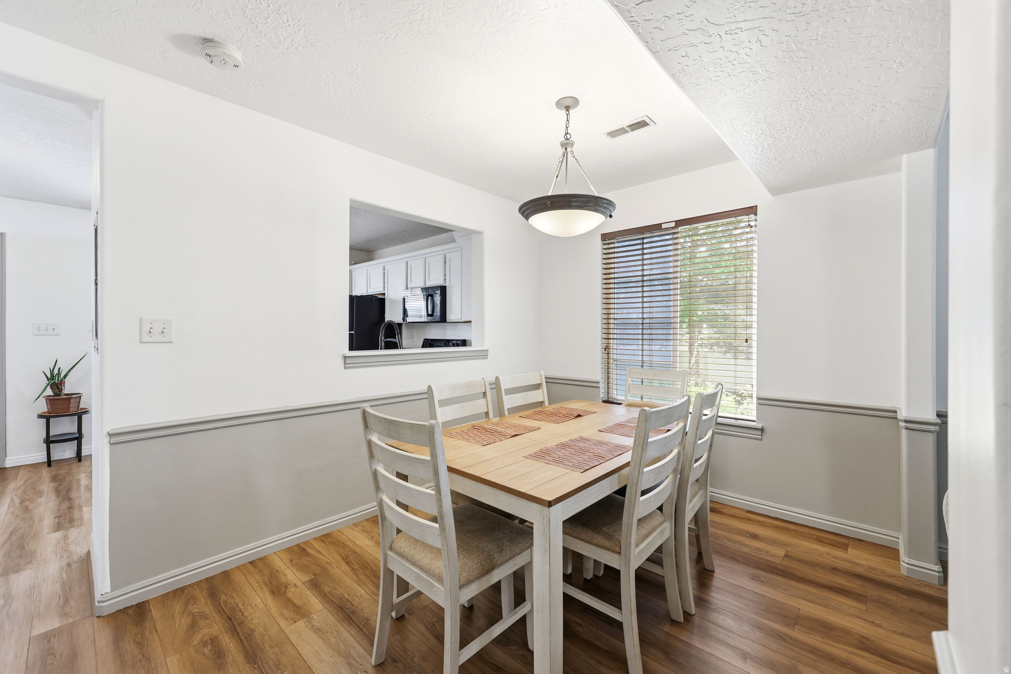 Dining area featuring wood-finish flooring, a contemporary ceiling light fixture, a window with blinds, and a pass-through opening to the kitchen