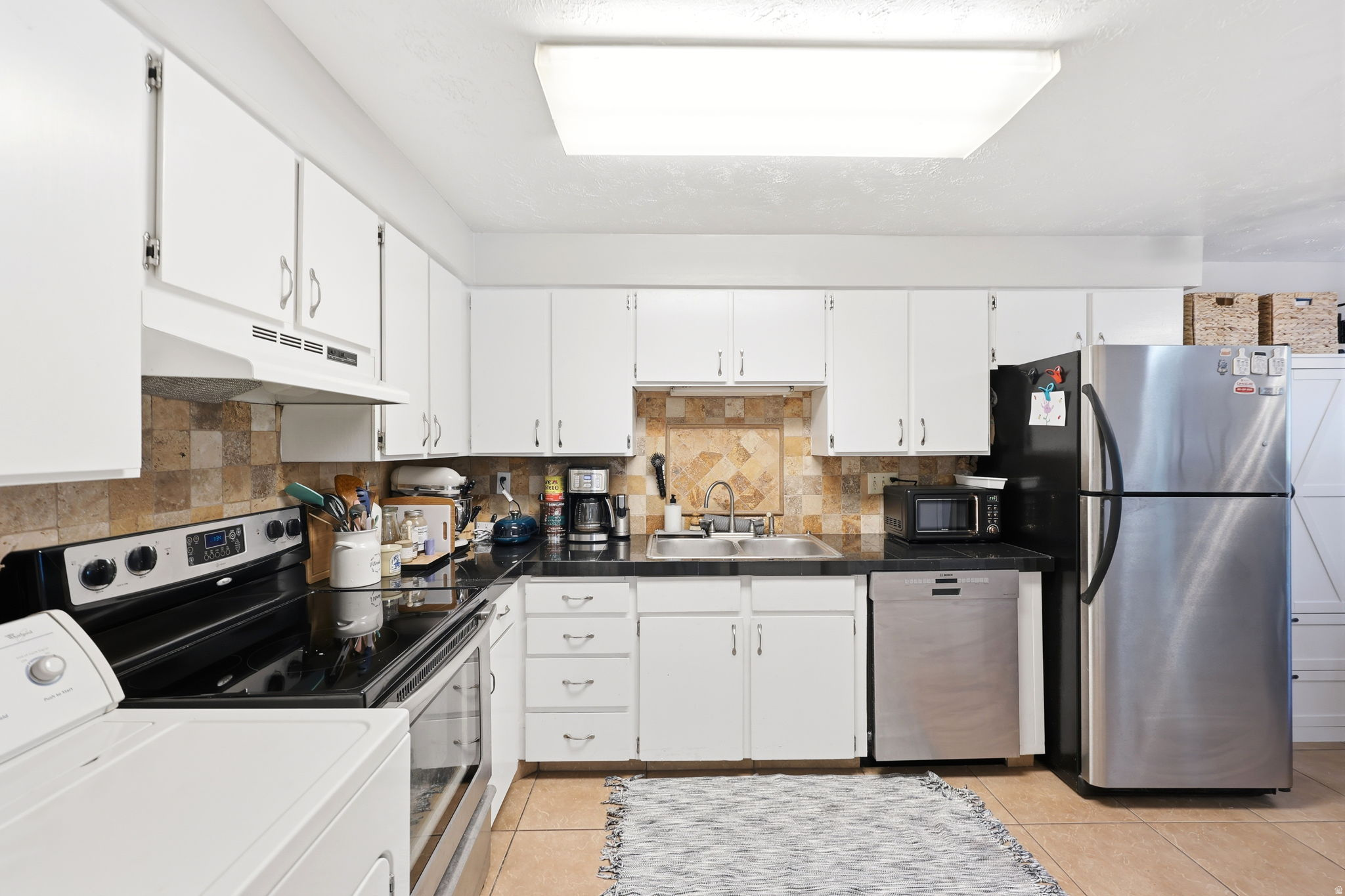 Kitchen featuring white cabinetry, black countertops, stainless steel refrigerator, stainless steel dishwasher, and tiled backsplash