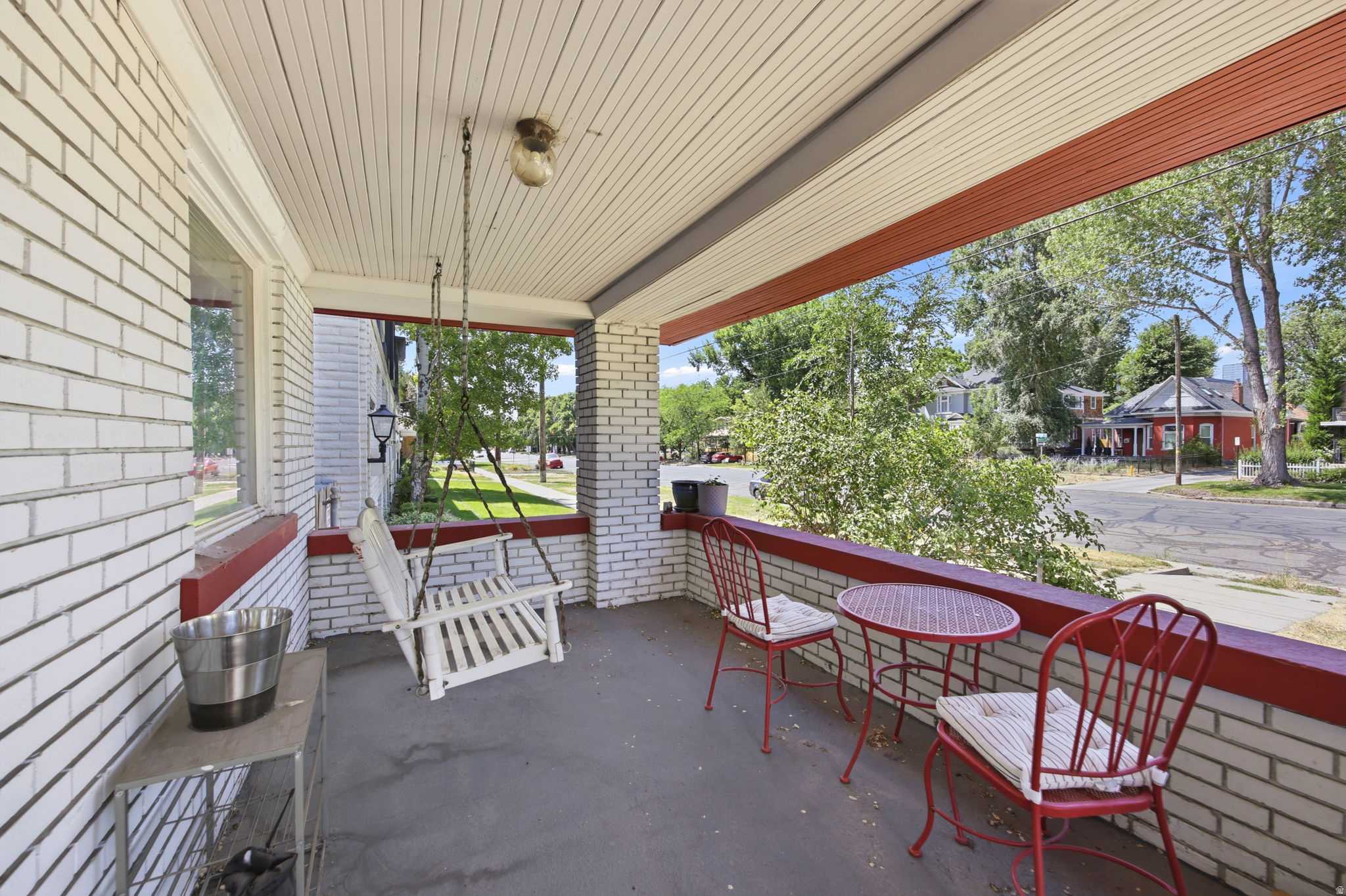 Covered front porch featuring white brick construction with red trim, a beadboard ceiling, a porch swing, and a concrete floor