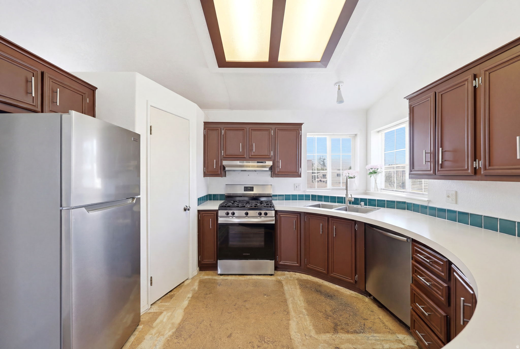 Kitchen featuring dark wood cabinetry, white countertops, stainless steel appliances, a gas range with an overhead hood, and a dual basin sink beneath a window