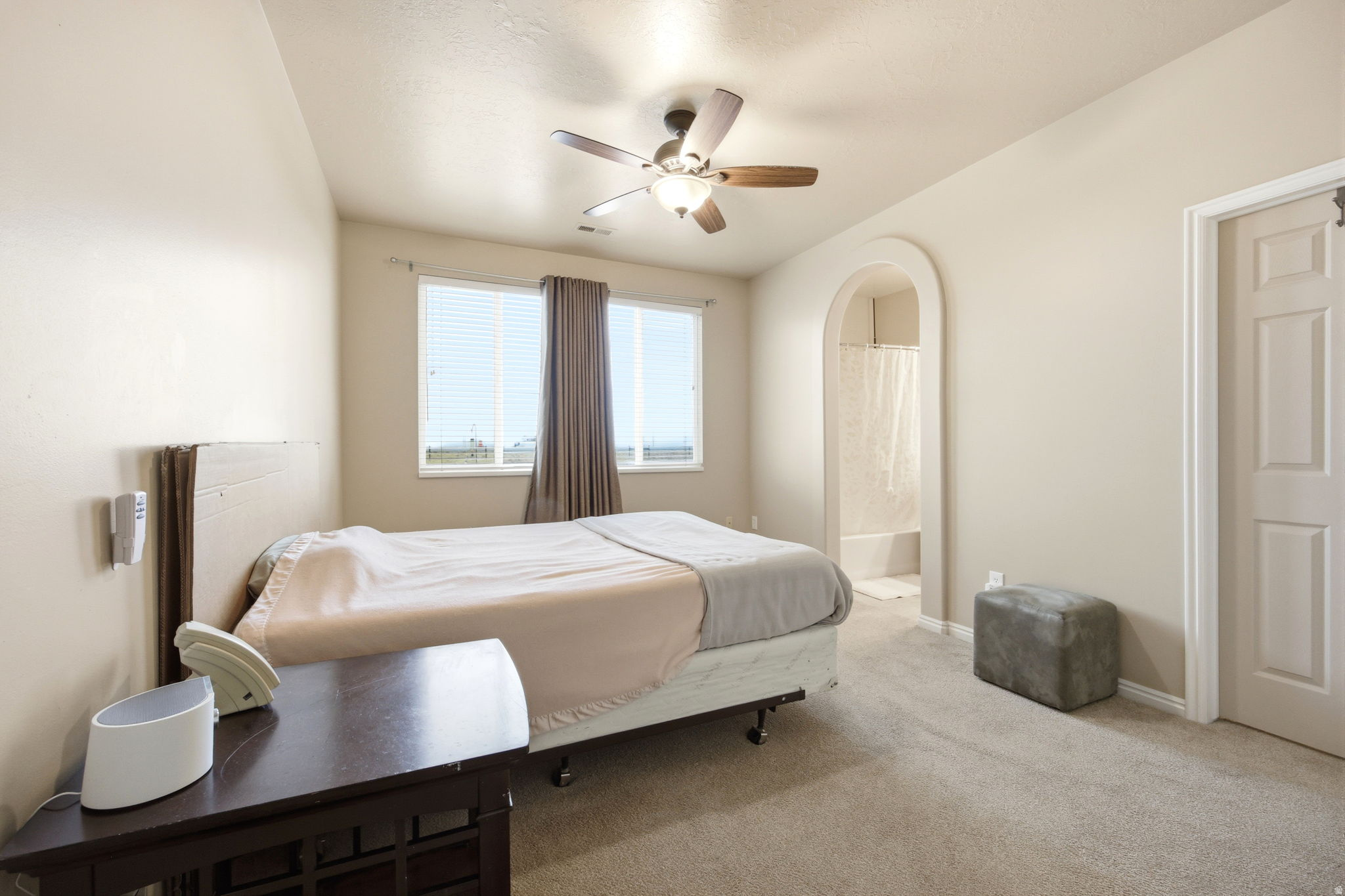 Carpeted bedroom with a ceiling fan light fixture, arched doorway to a bathroom, and a window with blinds and drapes