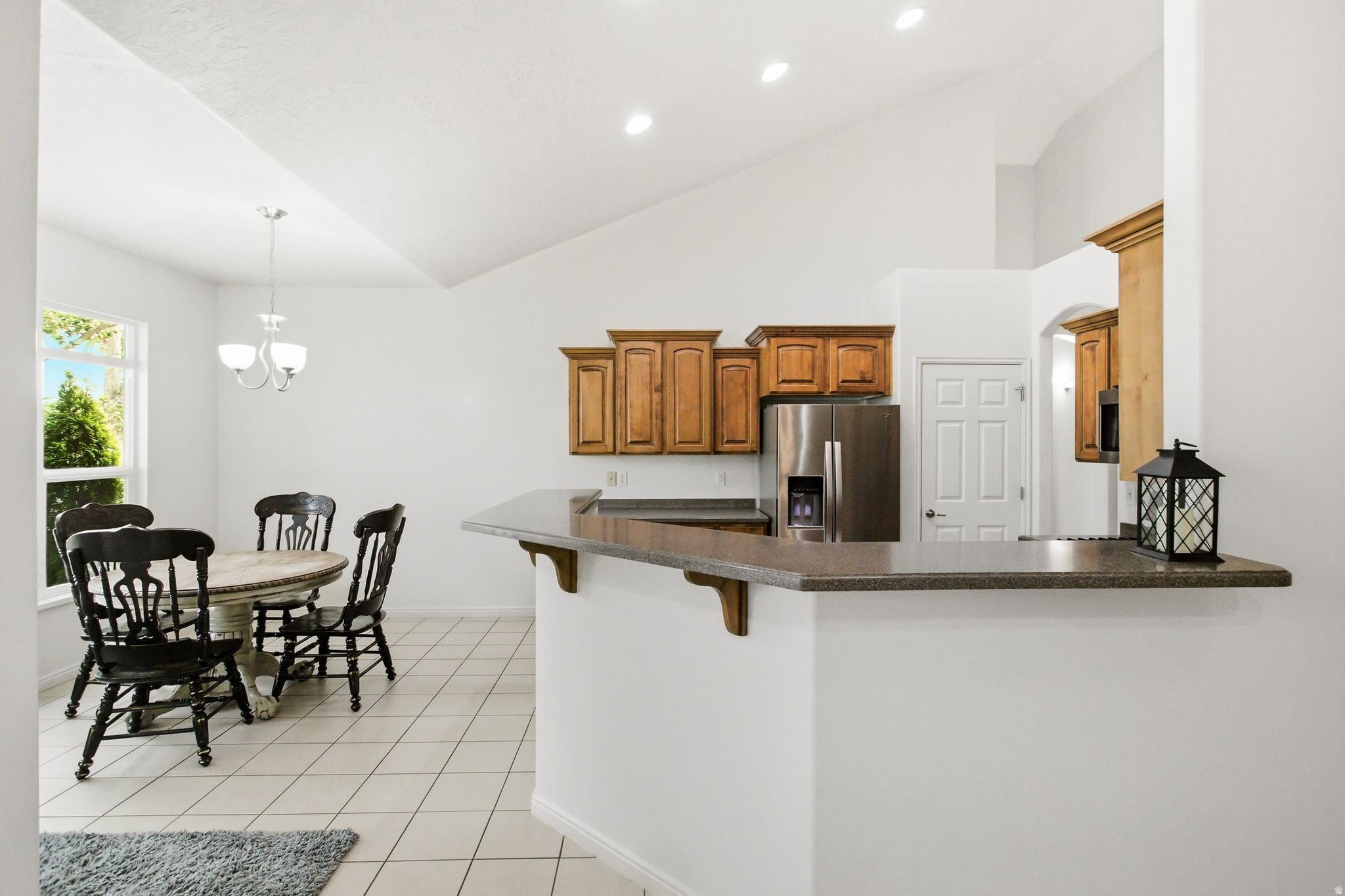 Open-concept kitchen and dining area featuring a vaulted ceiling, recessed lighting, wood cabinetry, stainless steel refrigerator, and a breakfast bar with a stone-finish countertop