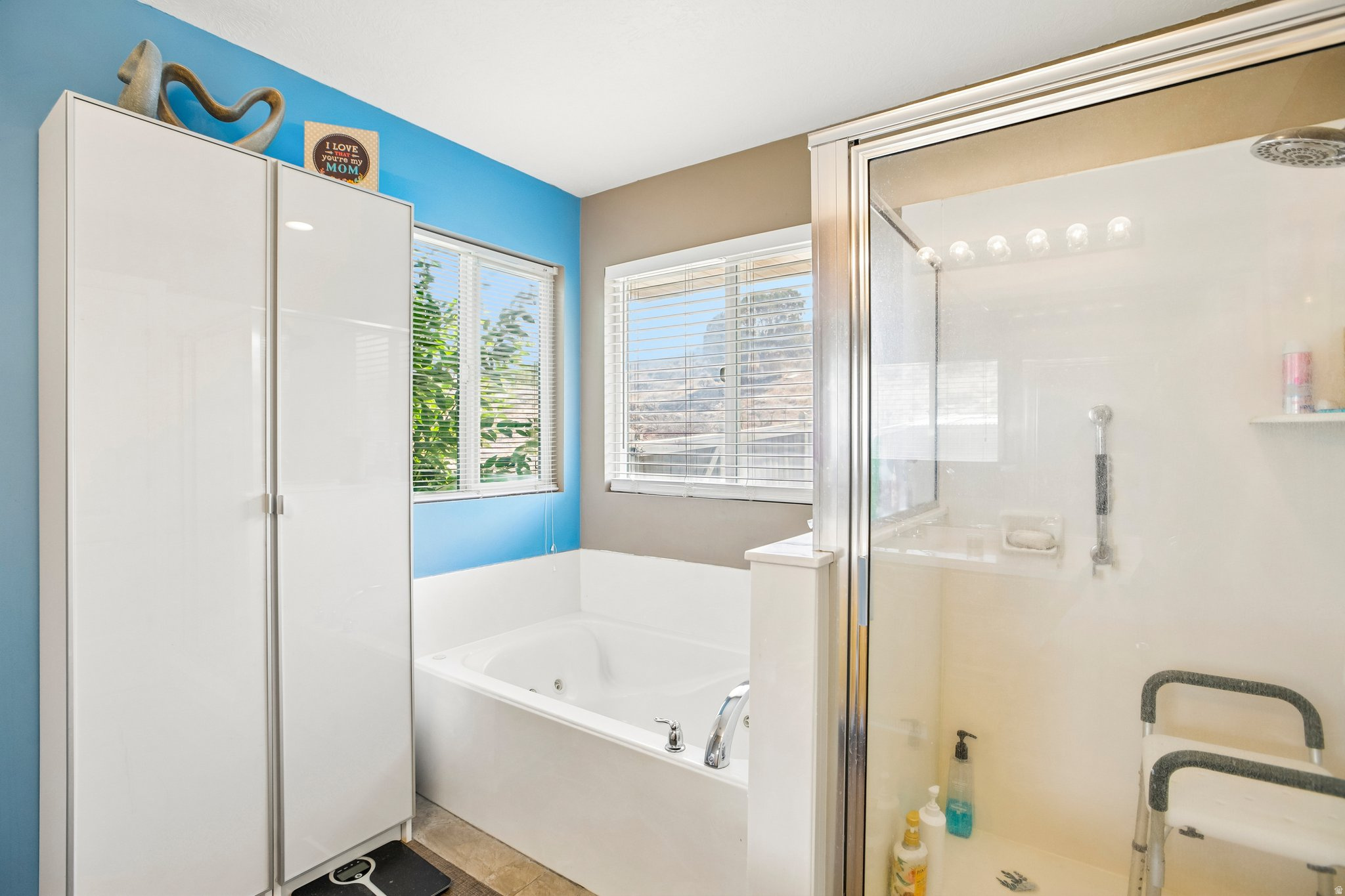 Bathroom featuring a built-in jetted tub, enclosed glass shower, dual windows with horizontal blinds, and two tall white storage cabinets