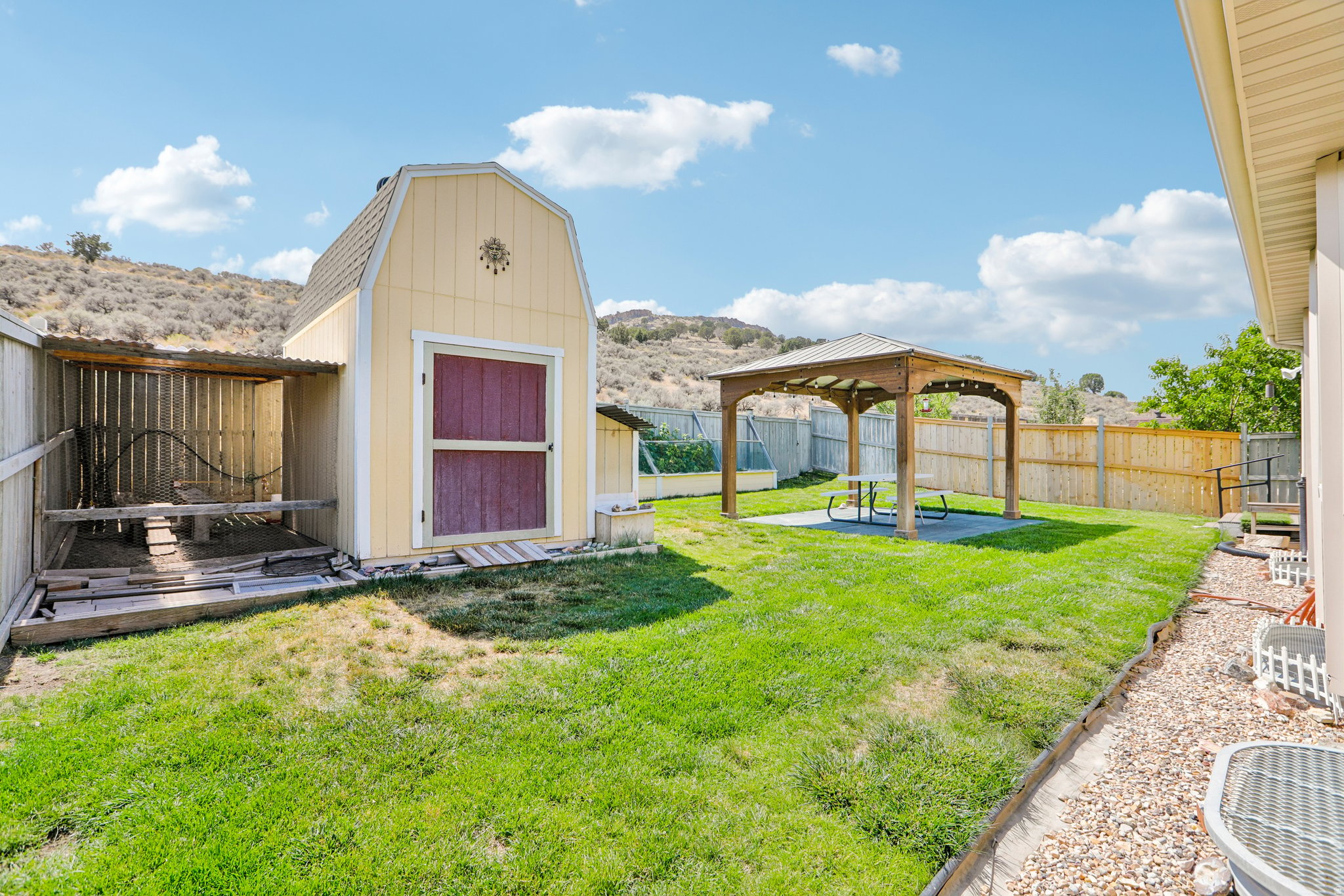 Expansive backyard featuring a lush lawn, wooden gazebo over a concrete patio, privacy fencing, a gambrel-roofed shed, and a covered animal enclosure