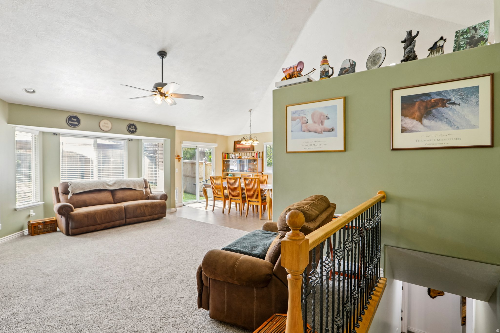 Open-concept living space featuring a bay window, carpet flooring, vaulted ceilings, a ceiling fan, and a wood-finish staircase with wrought iron balusters