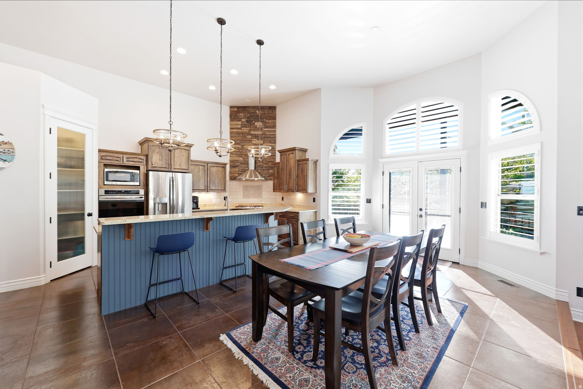 Spacious kitchen and dining area featuring vaulted ceilings, stainless steel appliances, a breakfast bar island with beadboard paneling, tiled flooring, and multiple arched windows with plantation shutters