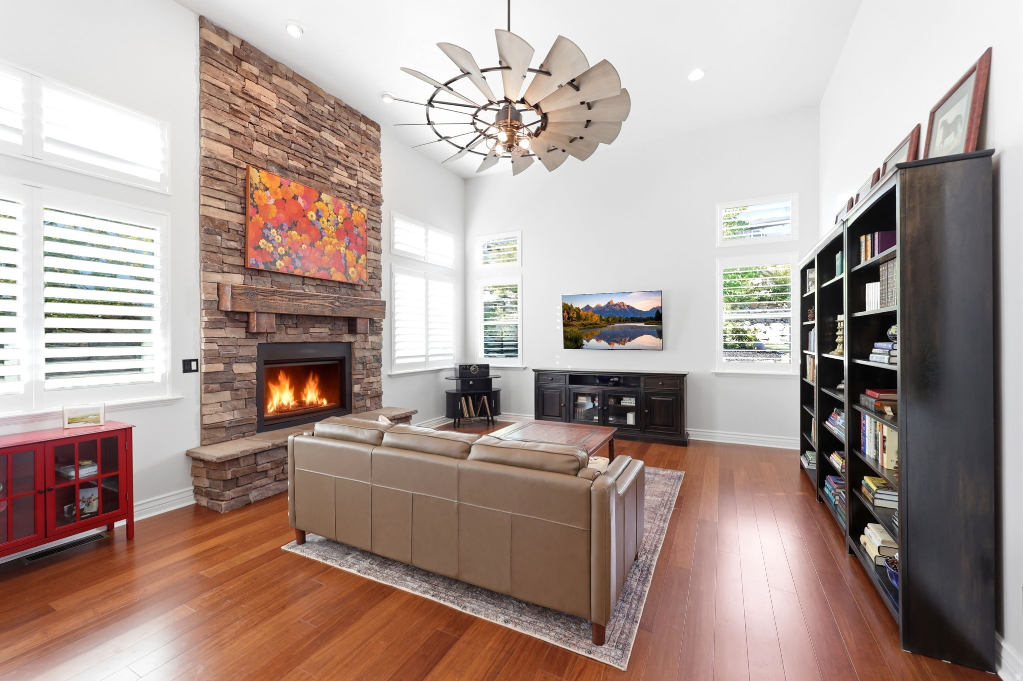 Spacious living area featuring a stone-clad fireplace with a substantial wood mantel, high vaulted ceilings, large plantation-shuttered windows, and wood-finish flooring