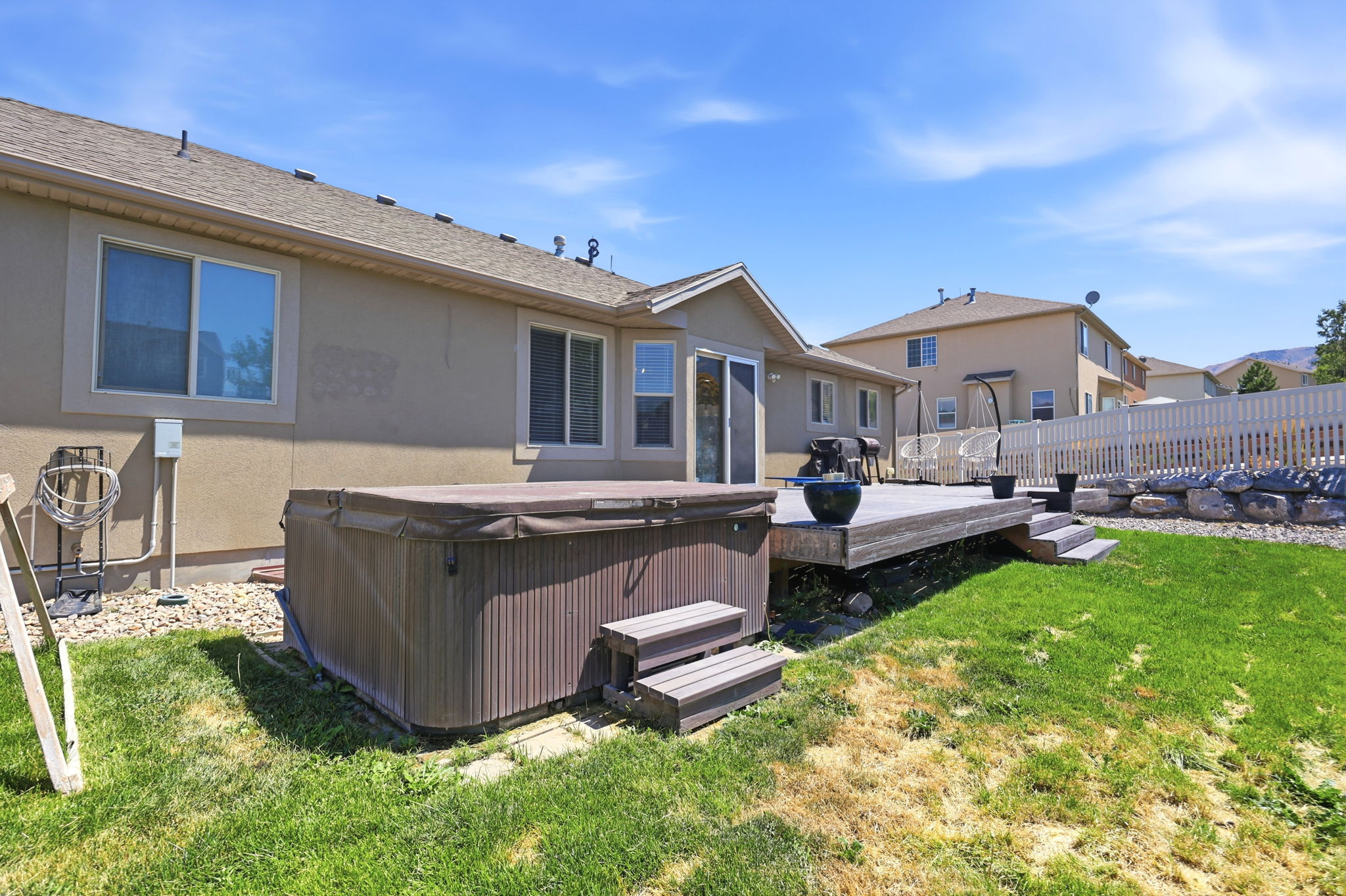 Rear exterior featuring a spacious wood deck, outdoor spa, sliding glass door, white picket fence, and a rock retaining wall