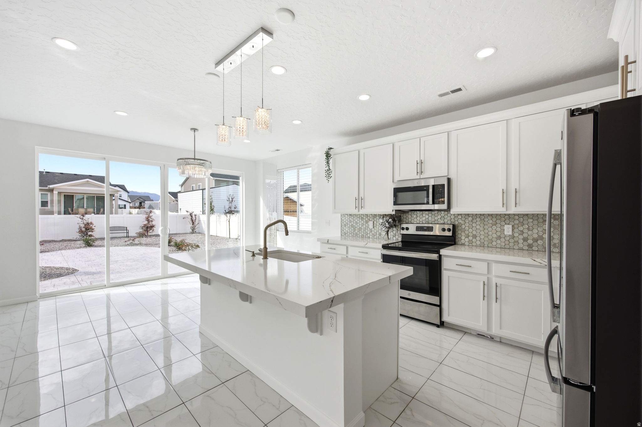 Open-concept kitchen featuring an island with a sink and modern pendant lighting, white cabinetry, integrated stainless-steel appliances, and a mosaic tile backsplash