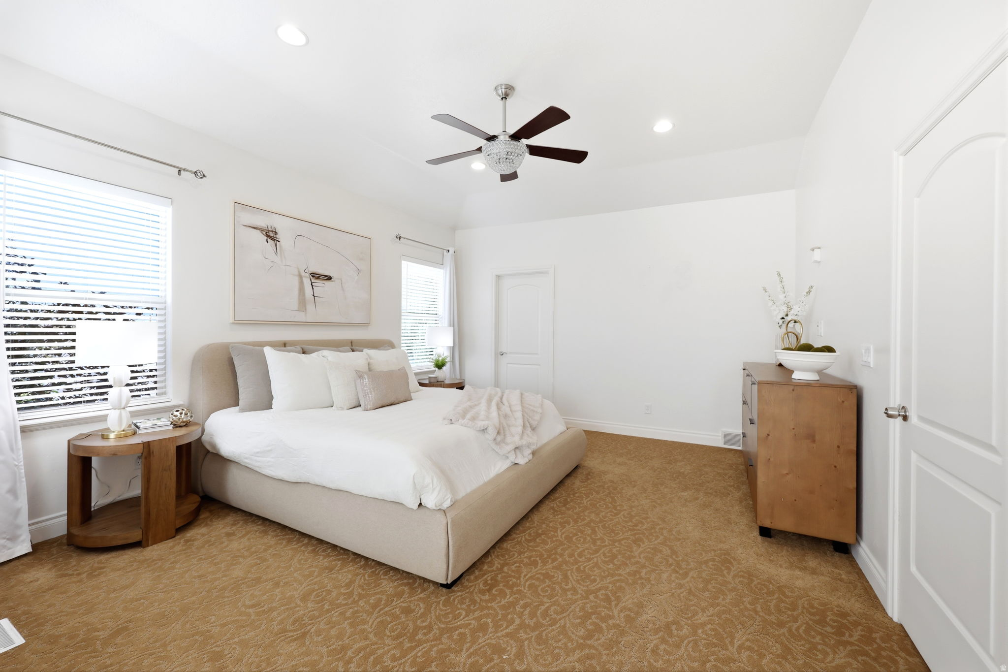 Spacious bedroom featuring light-colored walls, patterned carpeting, multiple windows with blinds, a ceiling fan with integrated lighting, and recessed lighting