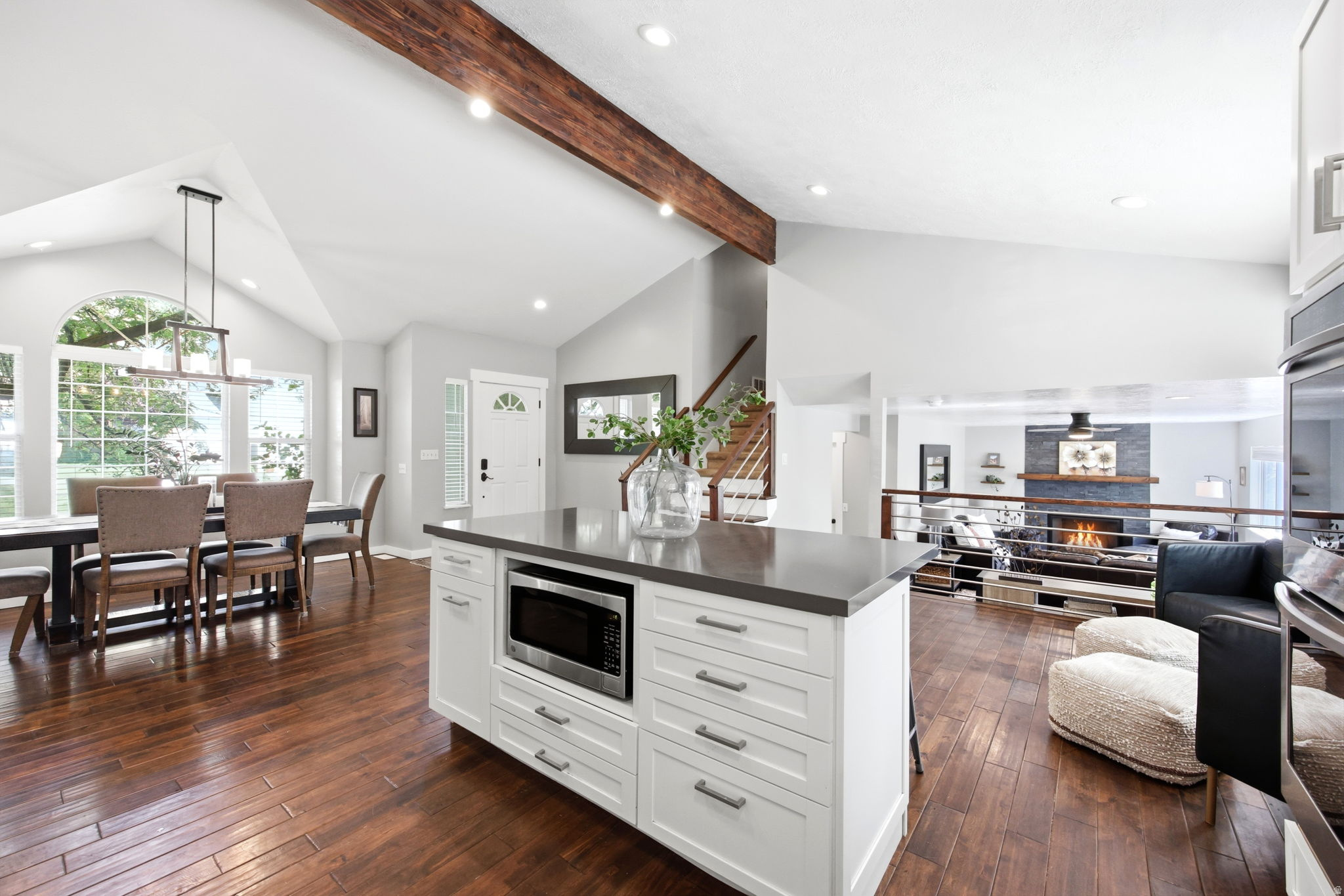 Open-concept living space featuring wood-finish flooring, a white kitchen island with a dark countertop and built-in microwave, vaulted ceilings with exposed wood beams, and a dining area with multiple windows