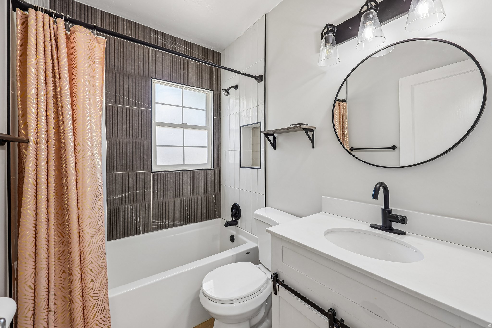 Modern bathroom featuring a ceramic vessel sink with matte black faucet and fixtures, white quartz countertop, circular framed mirror with chrome hardware, dual vanity lighting, and an enclosed shower-tub combination with textured tile surround and coordi