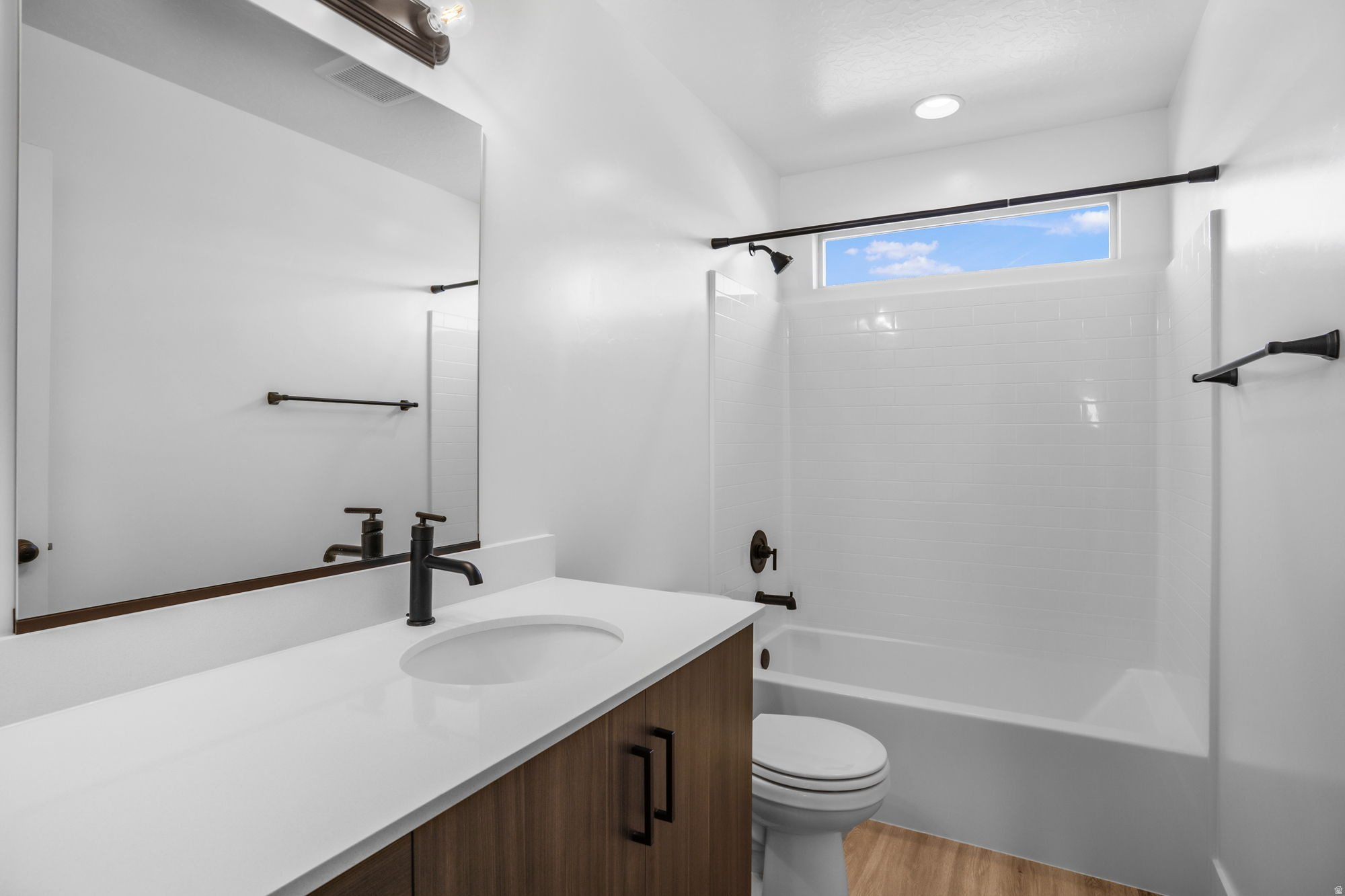 Bathroom featuring a wood-finish vanity with a white countertop and integrated sink, matte black fixtures, a large mirror, a tiled shower/tub combination, and a high horizontal window