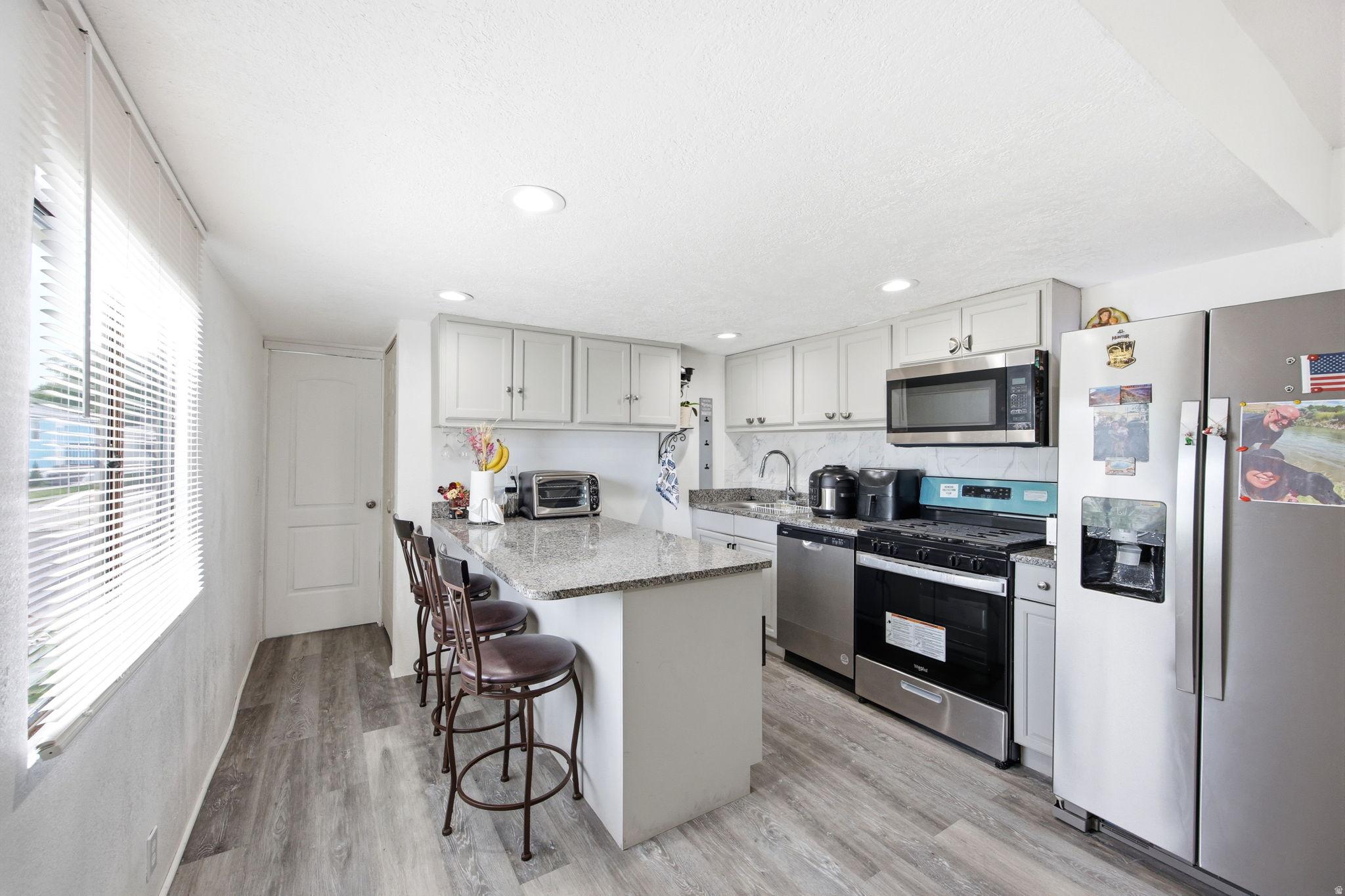 Contemporary kitchen featuring a granite-topped island with seating, stainless steel appliances, light gray cabinetry, and wood-finish flooring