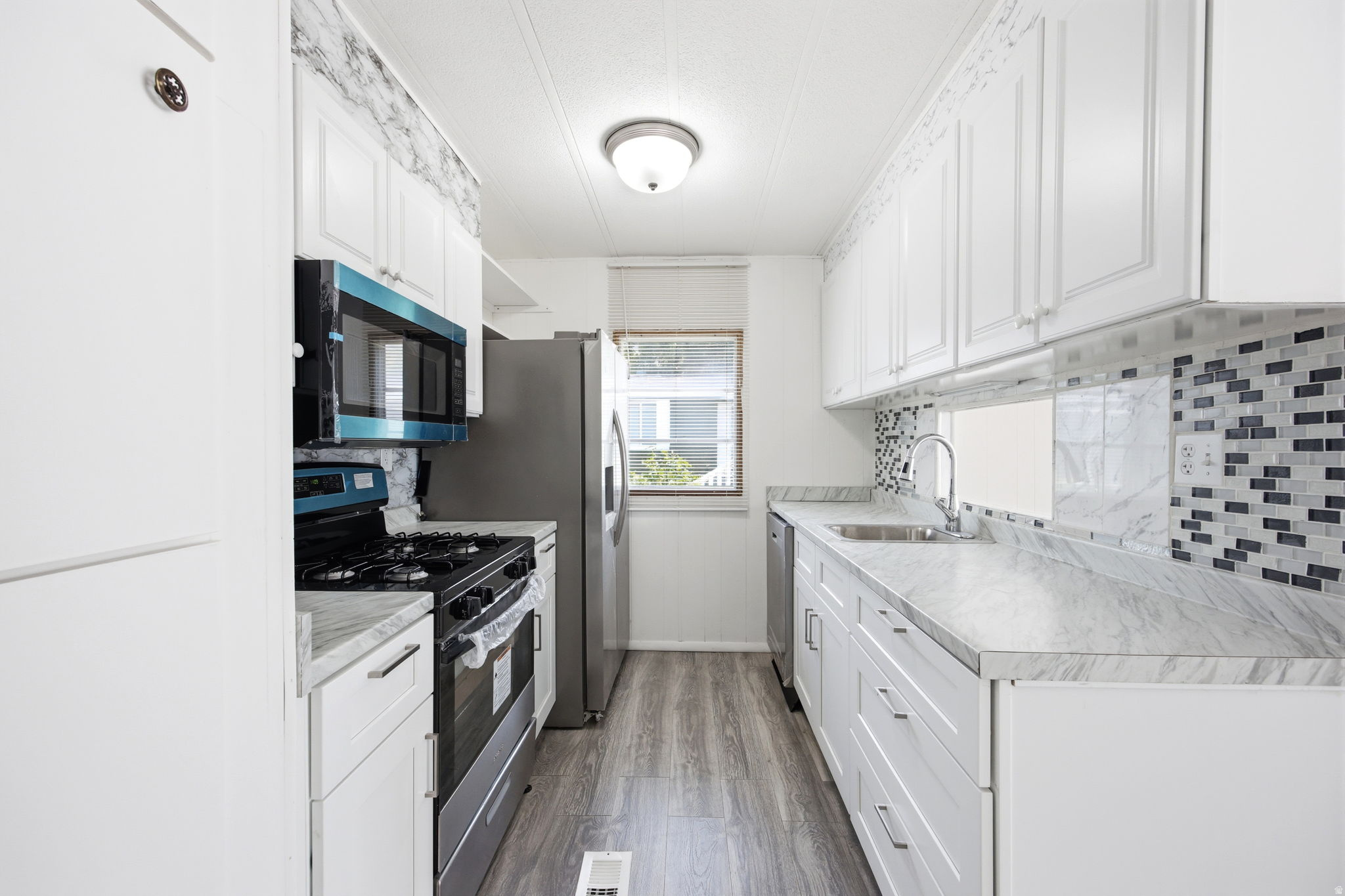 Galley kitchen featuring white cabinetry, wood-finish flooring, light-toned countertops, a mosaic tile backsplash, and a window providing natural light