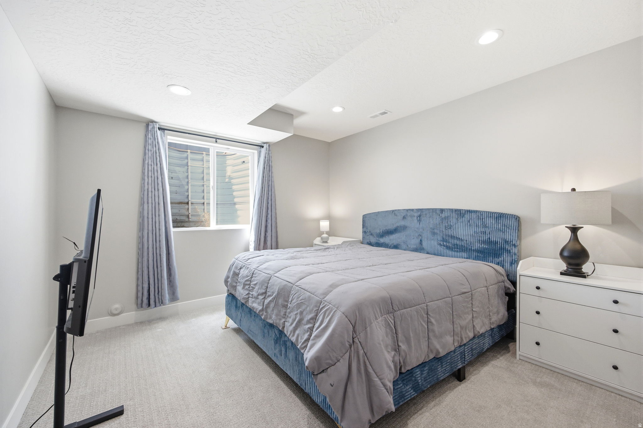 Spacious bedroom featuring light gray walls, recessed lighting, and a window with light-colored drapes