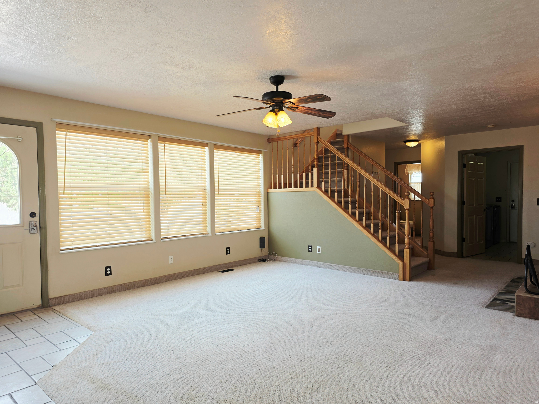 Carpeted living area with a wood staircase, three windows with blinds, a ceiling fan, and tile flooring at the entry