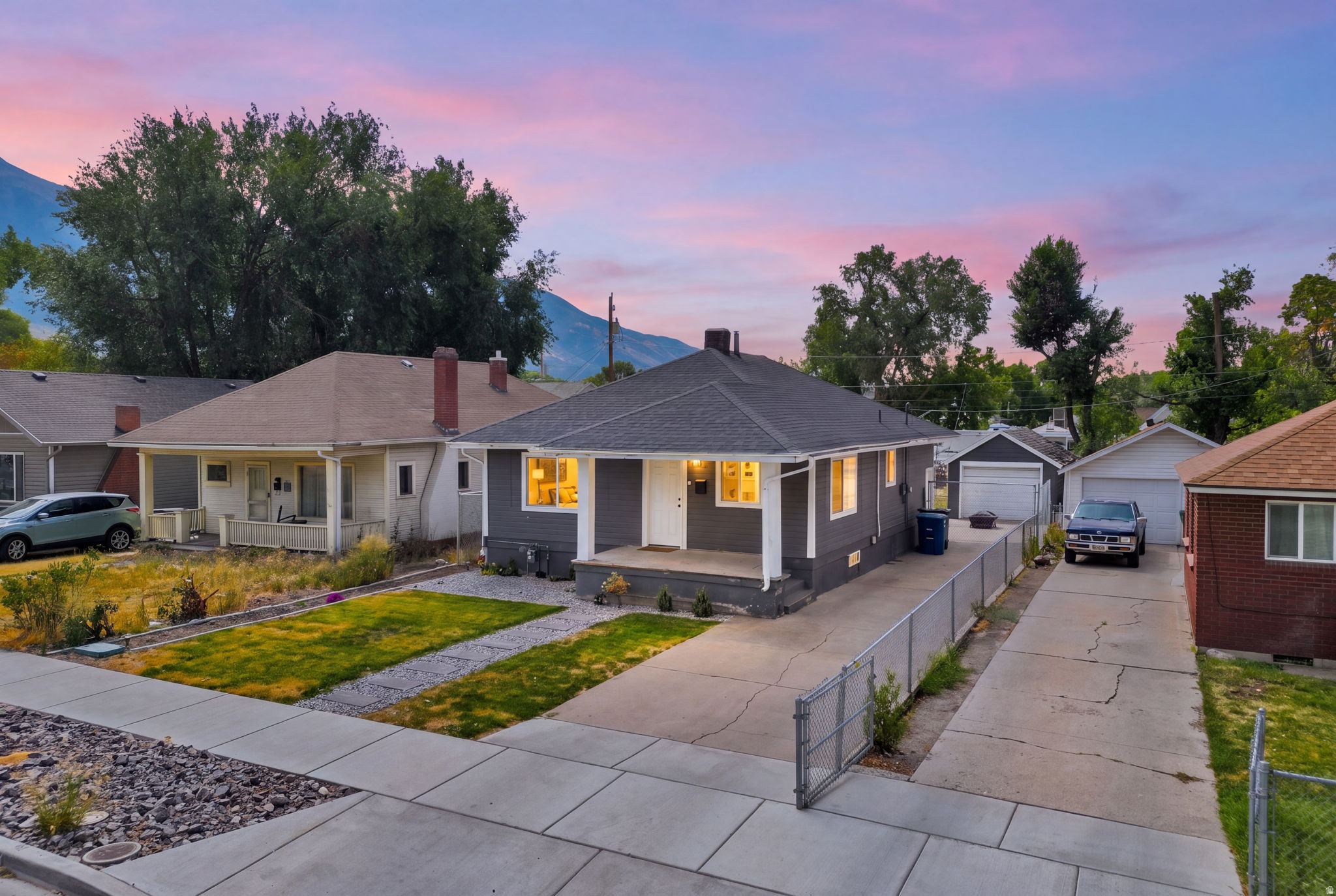Charming mid-century home featuring a freshly landscaped front yard with mature trees, modern concrete hardscaping, and illuminated interior spaces