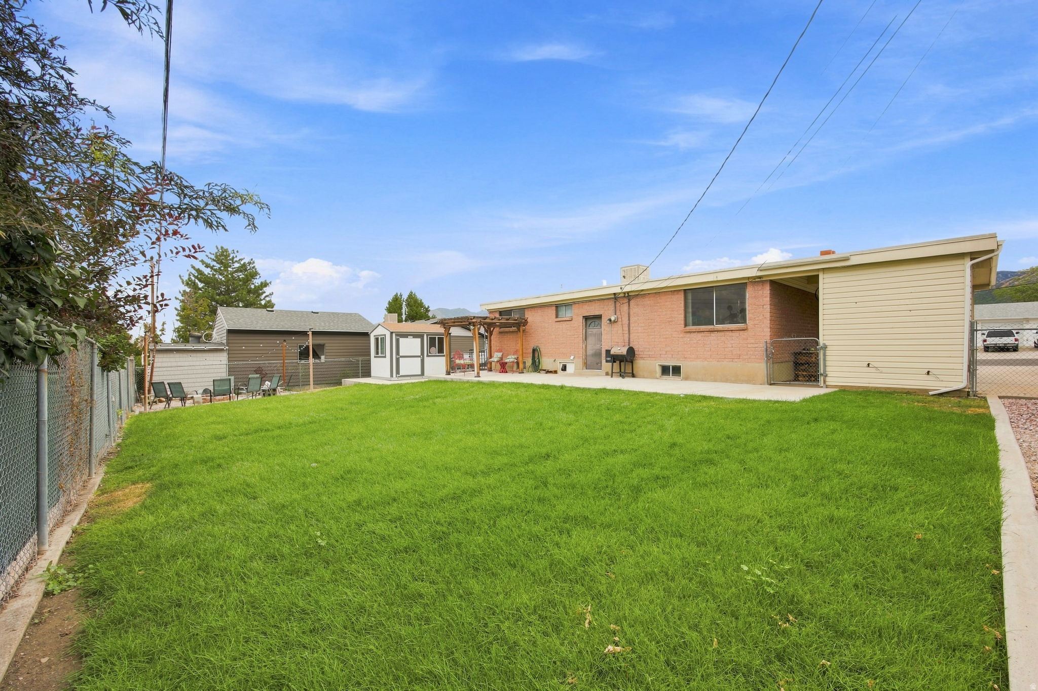 Expansive backyard featuring a manicured lawn, chain-link fencing, a detached storage shed, a brick exterior, and a covered patio area