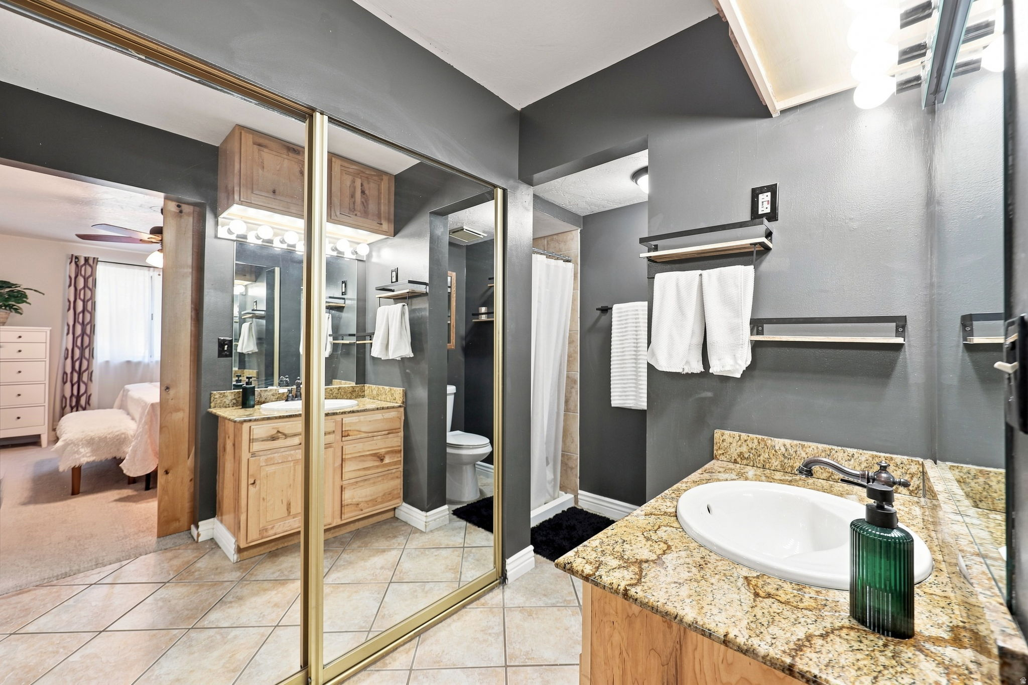 Bathroom featuring dual vanities with granite-style countertops, wood-finish cabinetry, ceramic tile flooring, and mirrored closet doors