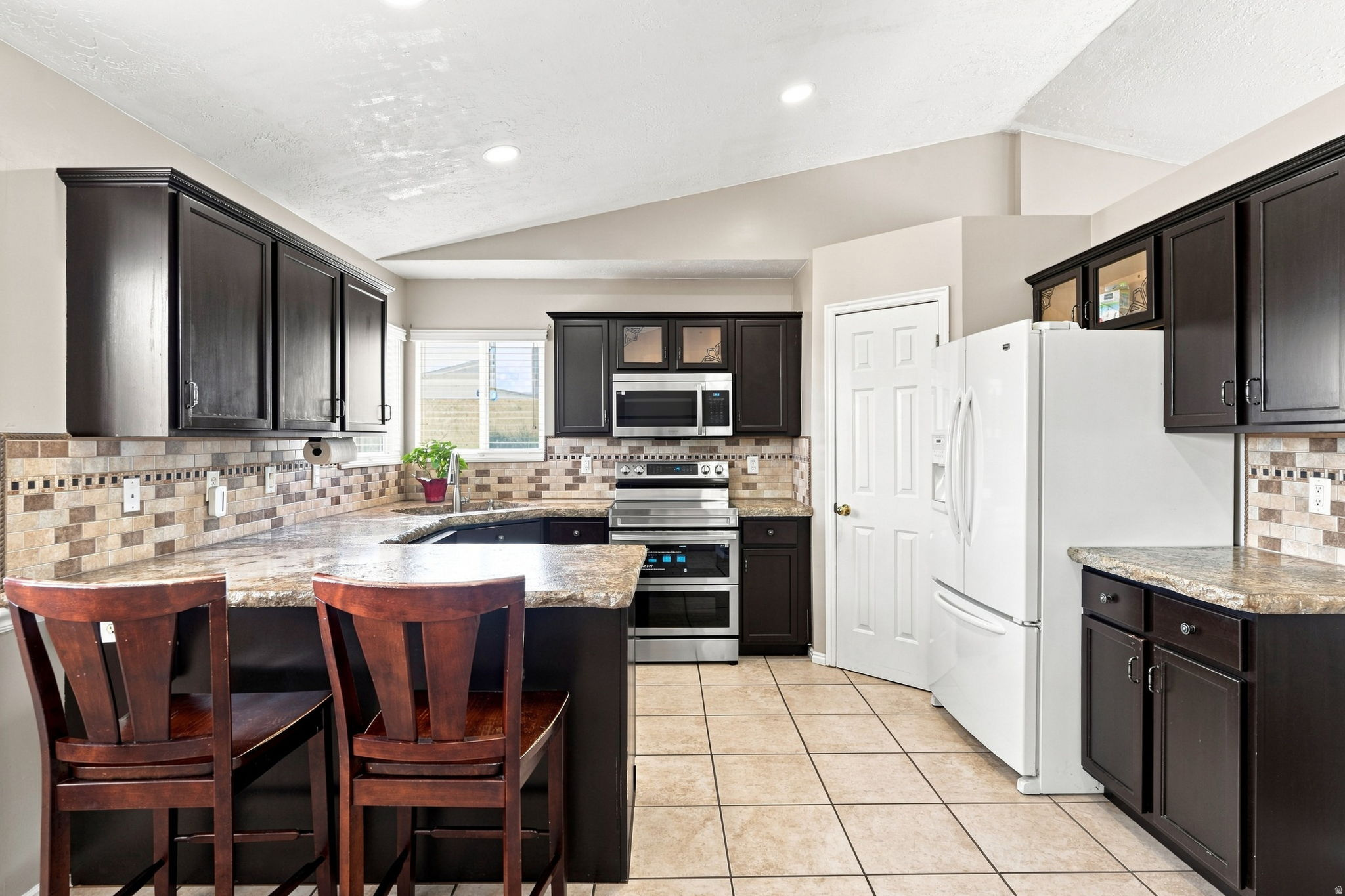 Kitchen featuring dark wood cabinetry, light-colored tile flooring, stone countertops, and a full tile backsplash