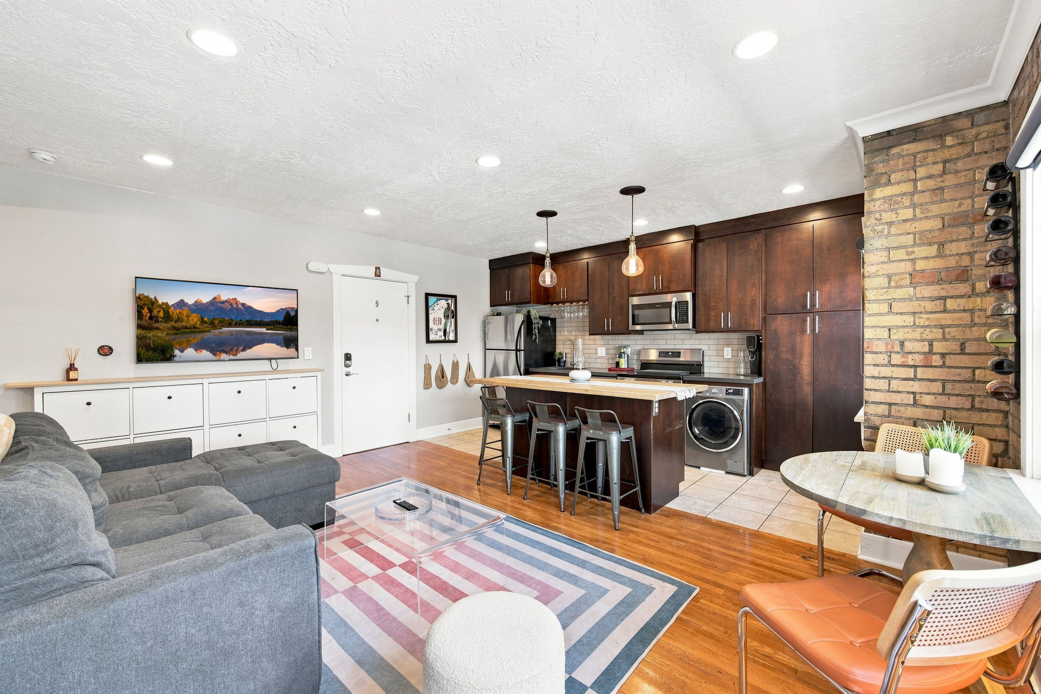Open-concept living area featuring hardwood flooring, recessed lighting, a brick accent wall, a kitchen island with pendant lighting, and dark wood cabinetry