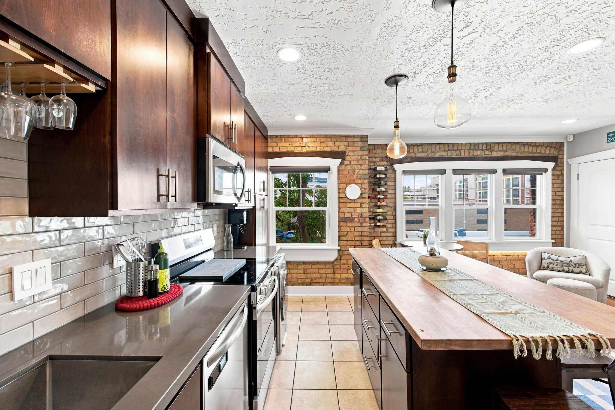 Kitchen featuring a wood-finish island, dark wood cabinetry, stainless steel appliances, a subway tile backsplash, and exposed brick accents