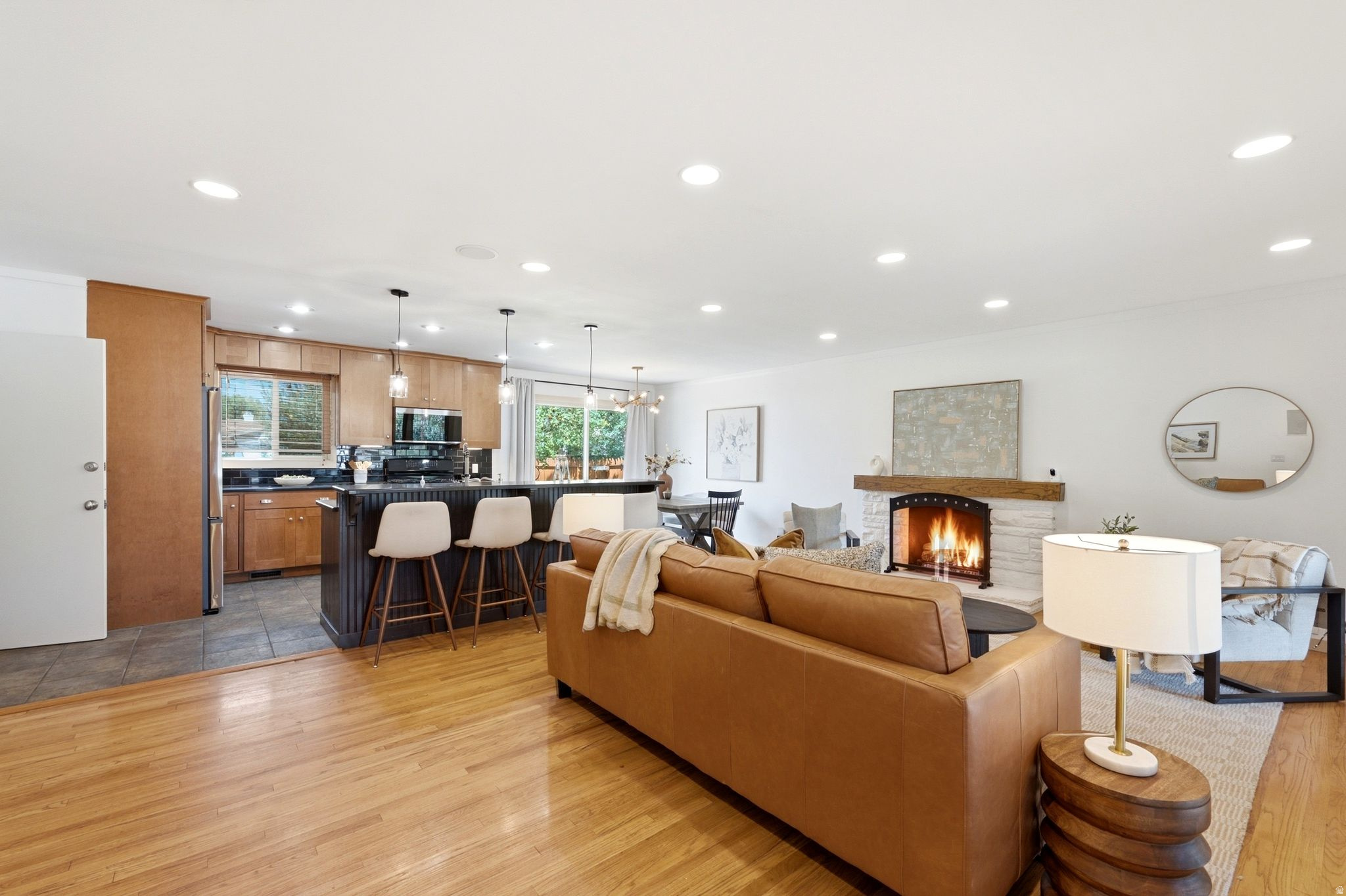 Hardwood flooring in the main living area, kitchen with wood cabinetry and dark countertops, central island with pendant lighting, stone-clad fireplace with wood mantel, and recessed ceiling lighting throughout