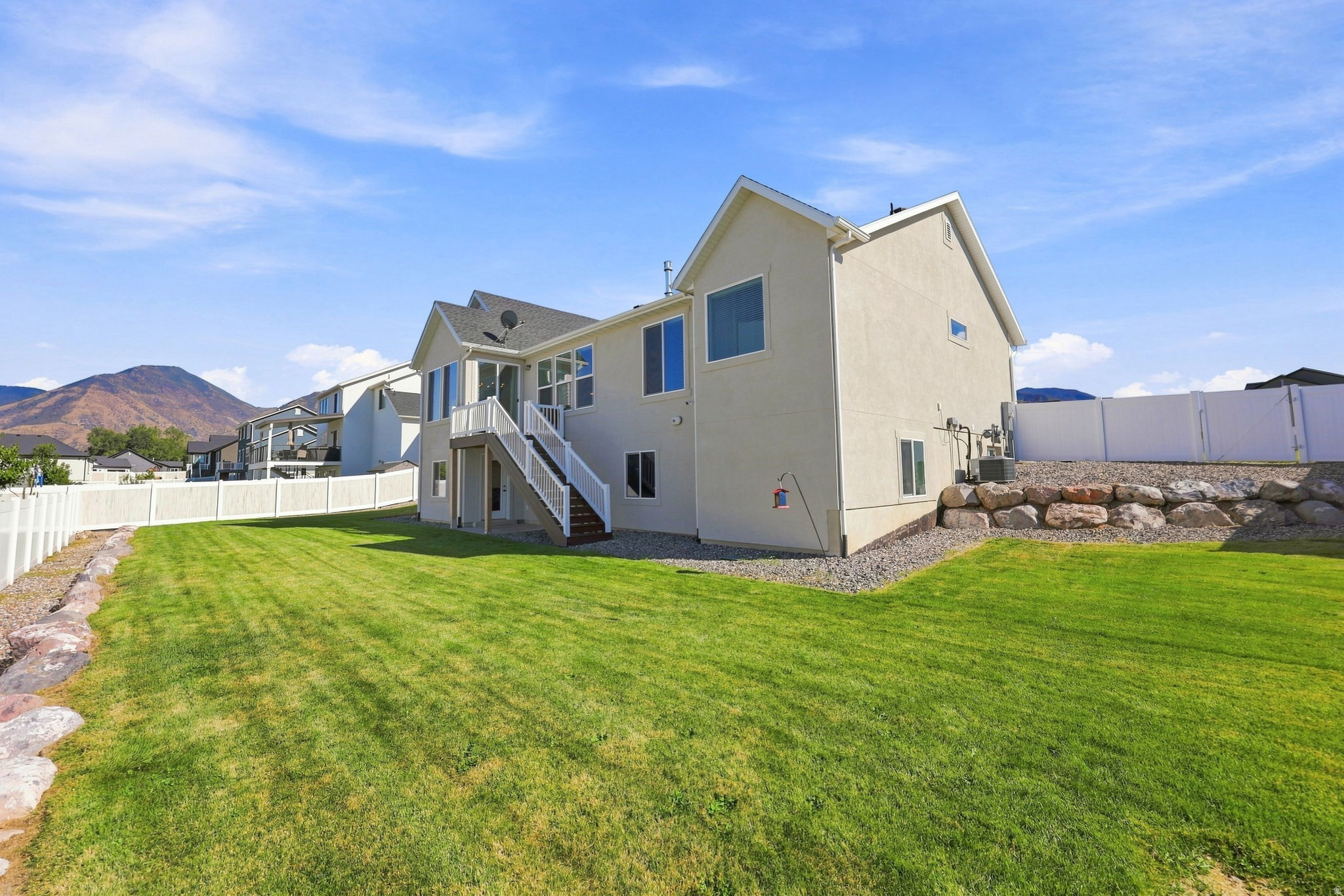 Expansive green lawn, white privacy fencing, multi-level deck with stairs, natural rock landscaping, and mountain backdrop