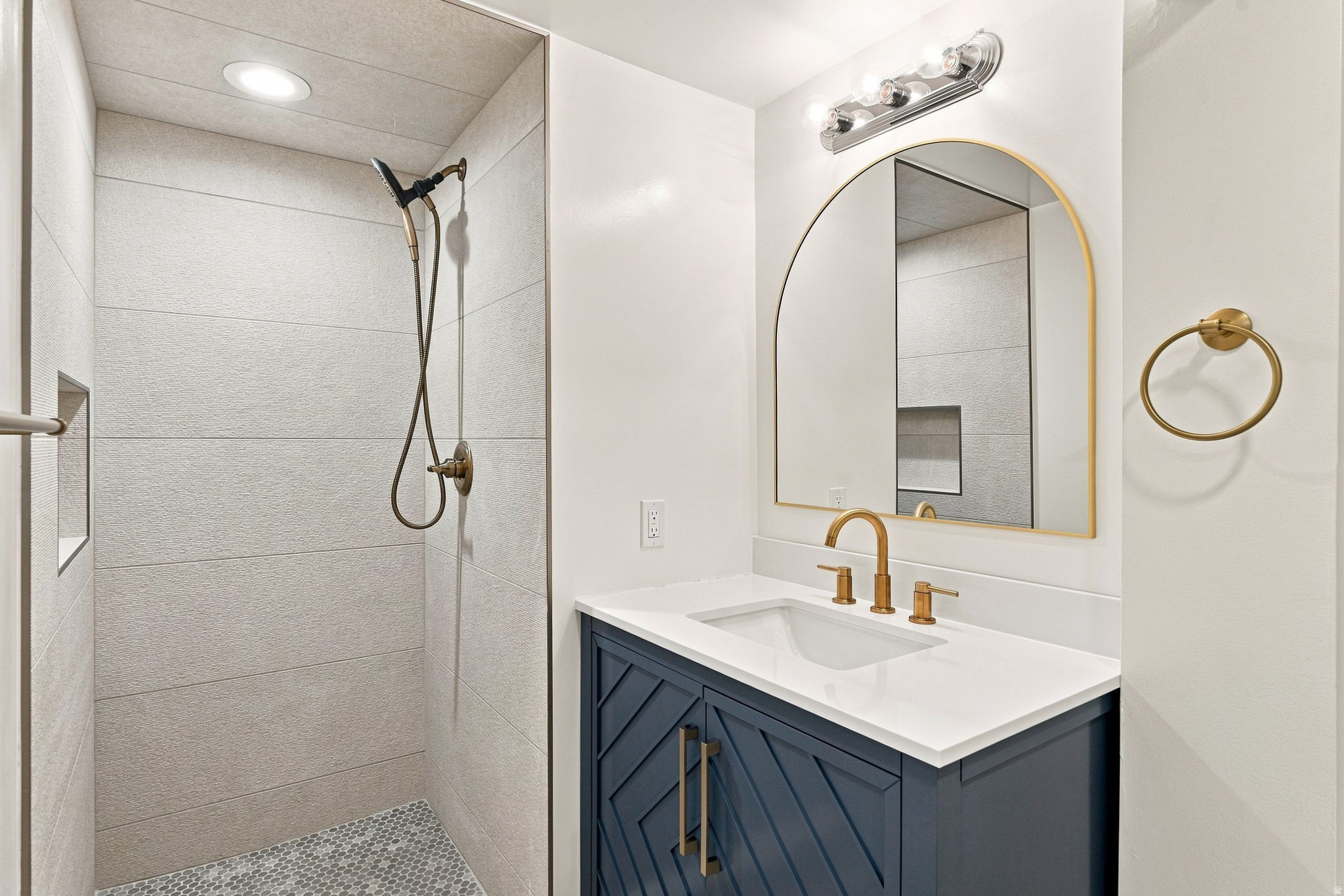 Contemporary bathroom featuring a navy blue vanity with gold hardware, a white countertop, an arched gold-framed mirror, and a tiled walk-in shower with a recessed niche