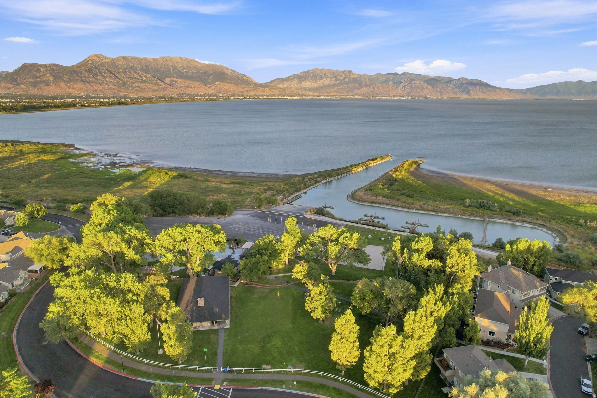 Aerial perspective of a property featuring a curved asphalt street, manicured lawn, and mature trees, with a lake and mountains in the distance
