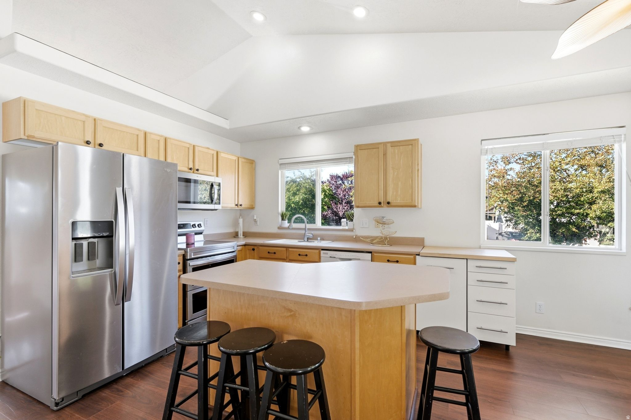 Kitchen featuring a vaulted ceiling, wood-finish flooring, light wood cabinetry, stainless steel appliances, and an island with seating