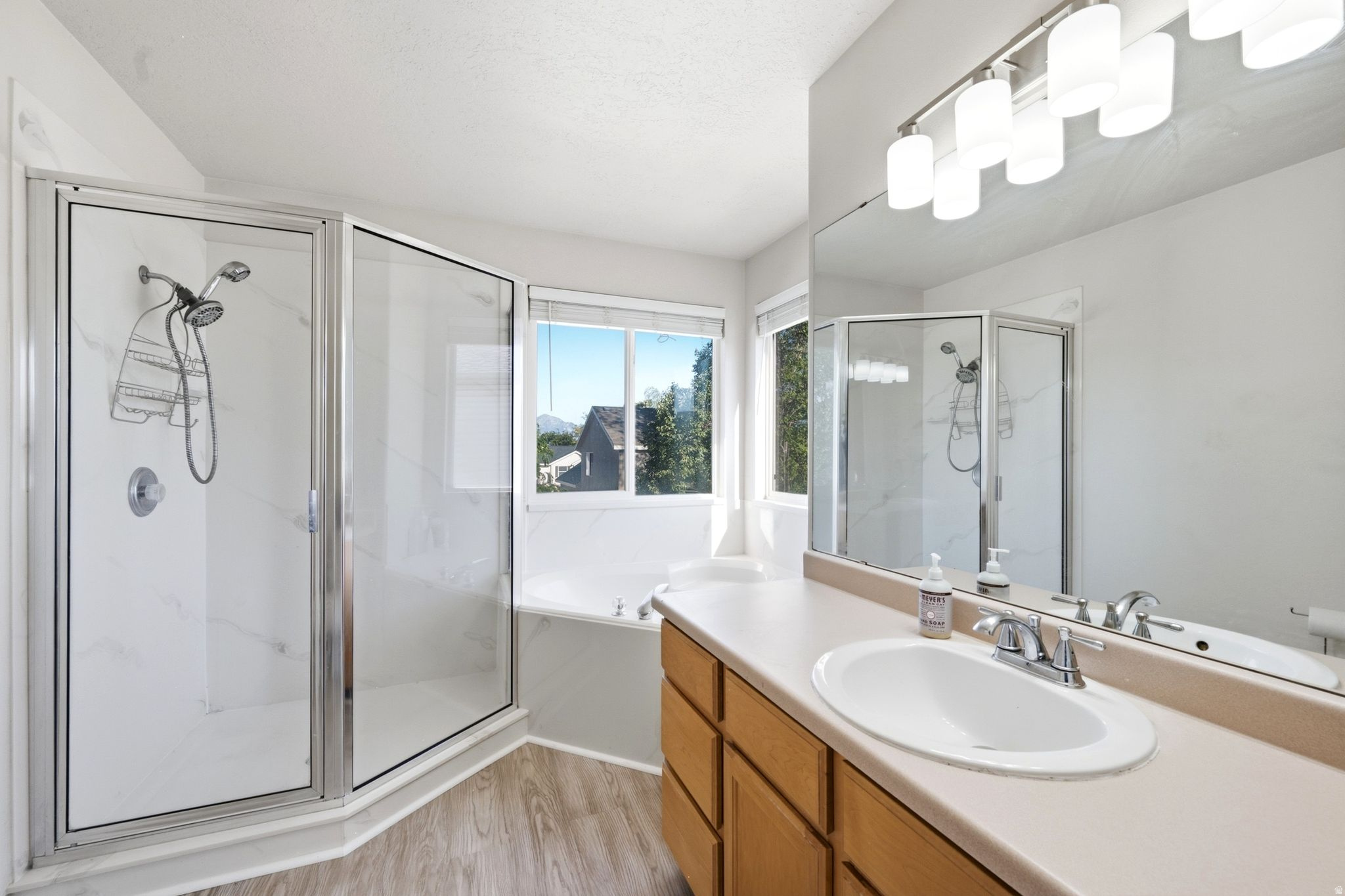 Walk-in shower with clear glass enclosure, built-in bathtub, dual-sink vanity with light-colored countertop, wood-finish cabinetry, and wood-finish flooring