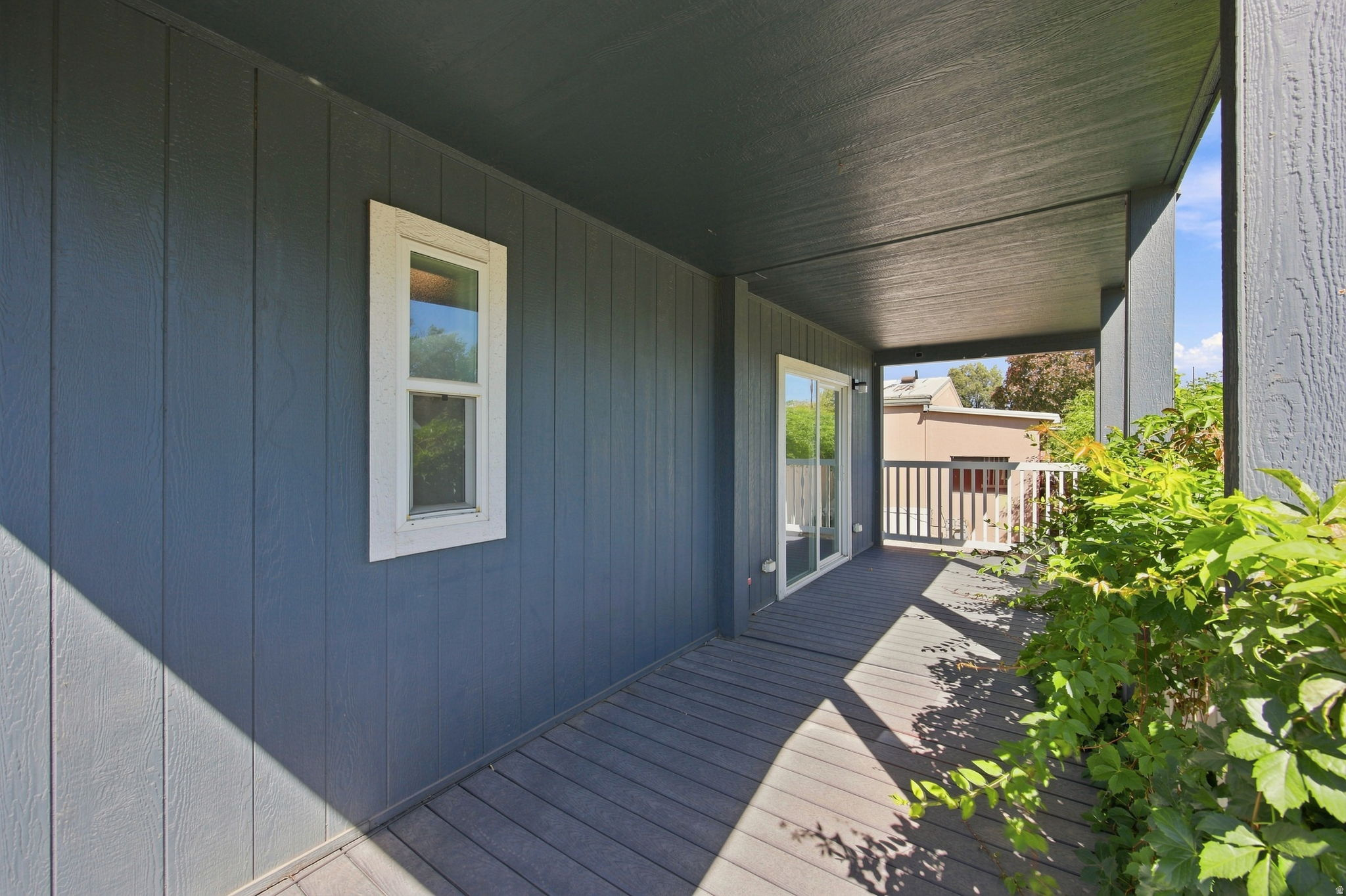 Covered outdoor deck with a sliding glass door, white window trim, and vertical panel siding