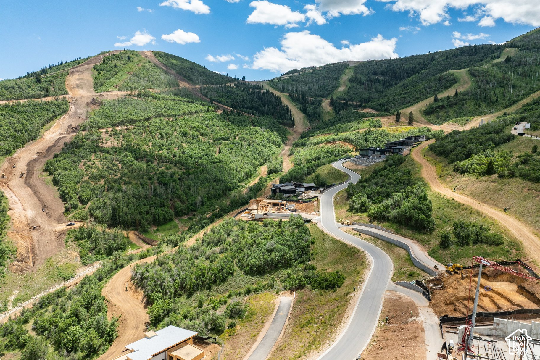 Aerial view of a mountain neighborhood under construction, with a winding road and modern homes among forested hills.