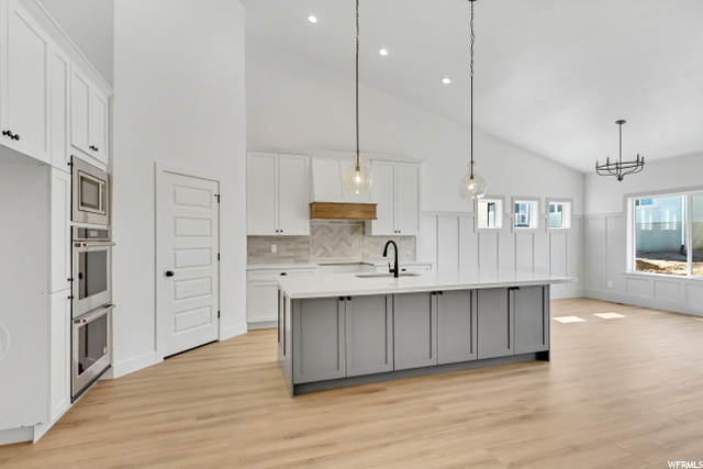kitchen with vaulted ceiling, stainless steel appliances including a double oven, light parquet floors, white cabinets, light countertops, pendant lighting, and an island with sink