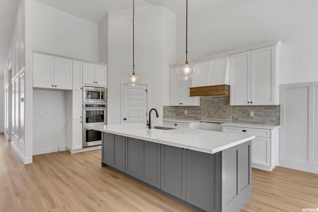 kitchen featuring stainless steel microwave, double oven, white cabinetry, light hardwood floors, light countertops, a kitchen island with sink, and pendant lighting