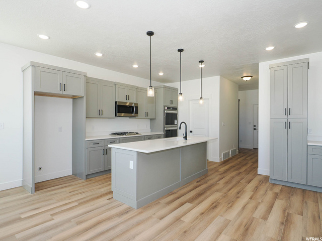 kitchen featuring double oven, microwave, white cabinets, pendant lighting, light hardwood flooring, a kitchen island with sink, and light countertops