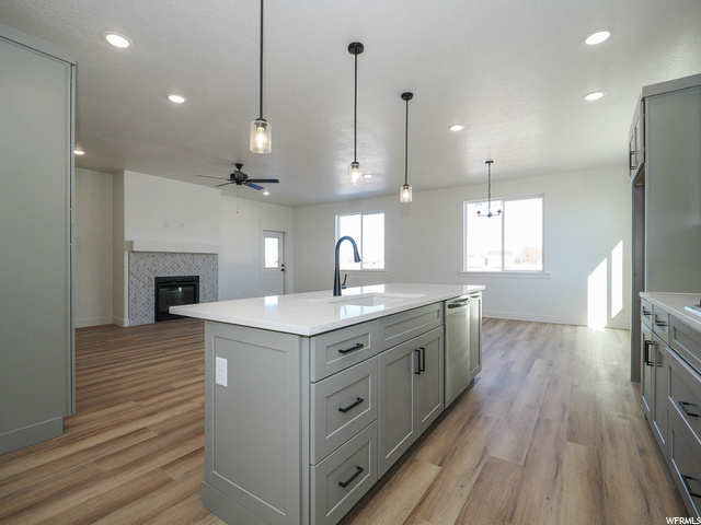 kitchen featuring natural light, a ceiling fan, a fireplace, light countertops, light hardwood floors, kitchen island sink, and pendant lighting