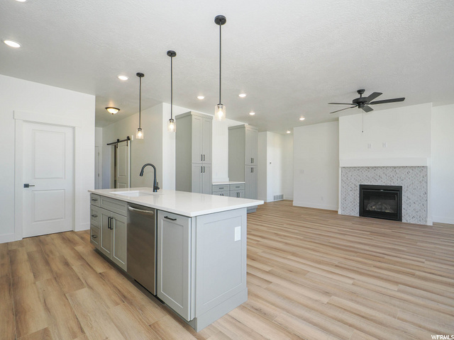 kitchen featuring a fireplace, a ceiling fan, dishwasher, pendant lighting, light countertops, kitchen island sink, and light hardwood flooring
