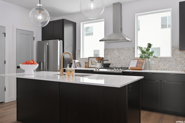 kitchen featuring a healthy amount of sunlight, gas stovetop, exhaust hood, stainless steel refrigerator, pendant lighting, dark brown cabinets, kitchen island sink, light countertops, and light hardwood flooring