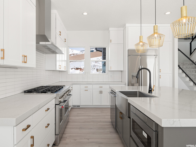 kitchen featuring refrigerator, gas range oven, exhaust hood, microwave, dishwasher, light countertops, pendant lighting, white cabinetry, and light hardwood flooring